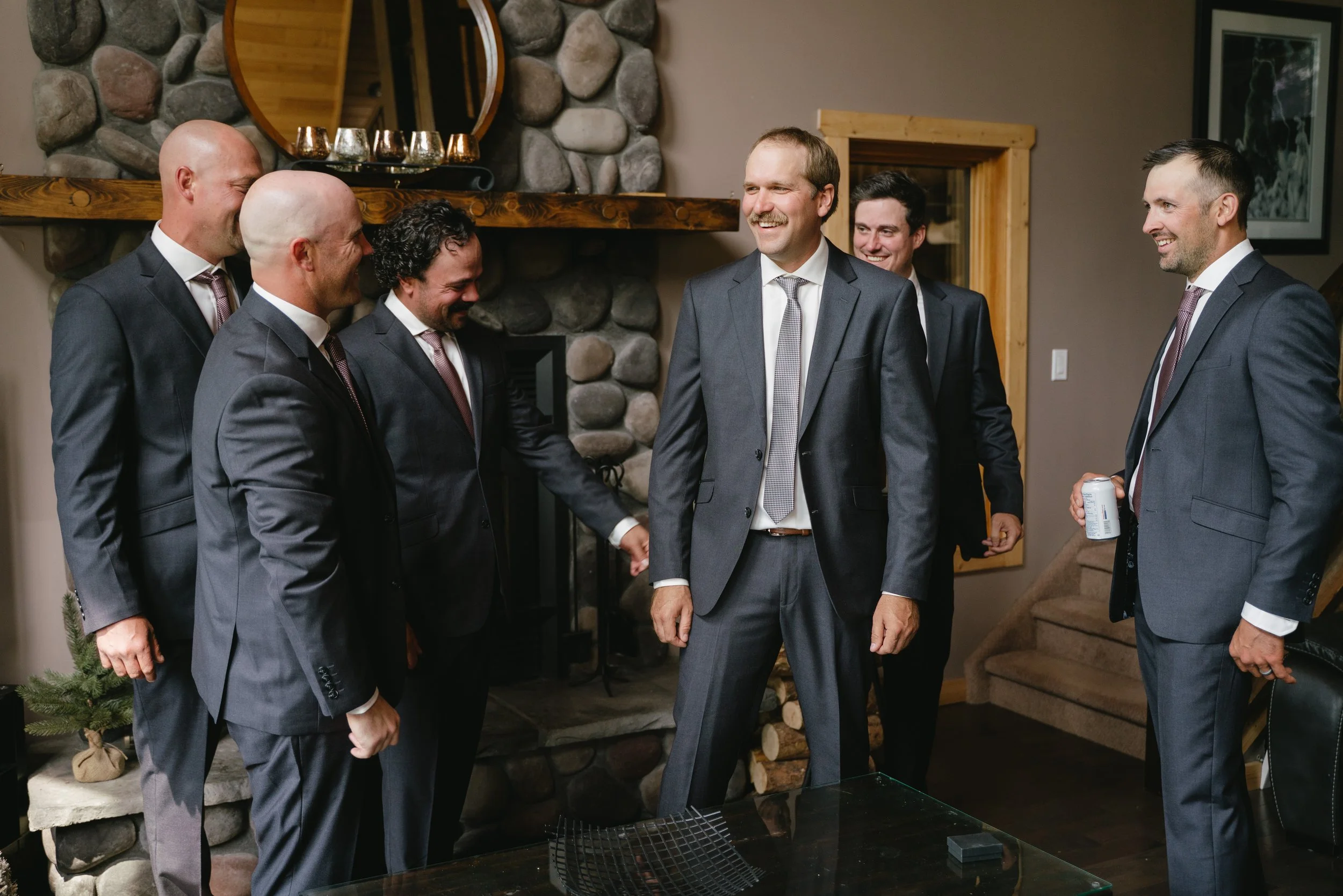 A group of six men dressed in suits are standing together inside a cozy room, smiling and enjoying themselves. They are engaged in conversation, with some holding drinks, and appear to be at a social gathering or celebration.