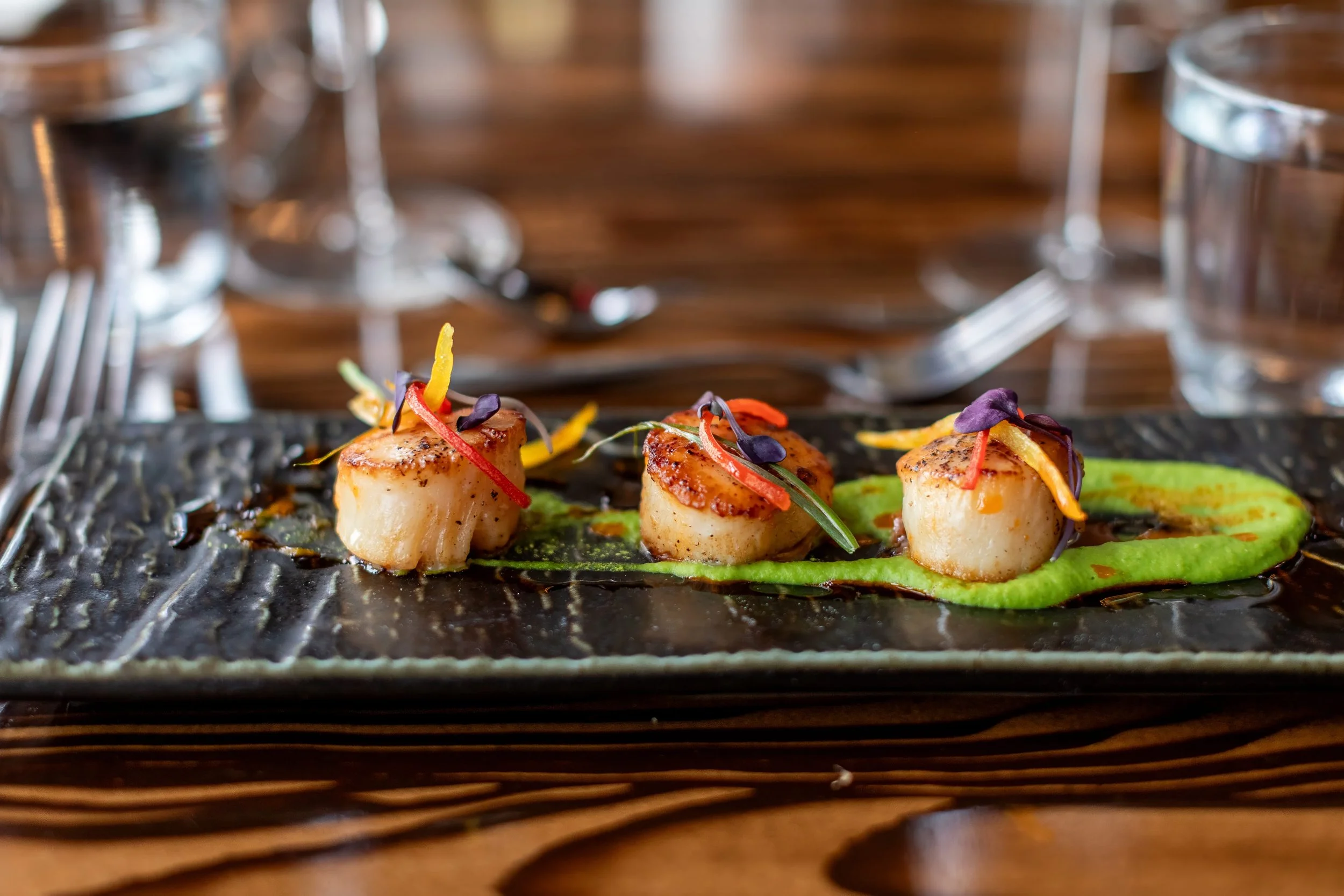 Three seared scallops garnished with microgreens and thin strips of colorful vegetables, served on a black textured plate with a green sauce underneath, set on a wooden table with drinking glasses and silverware in the background.