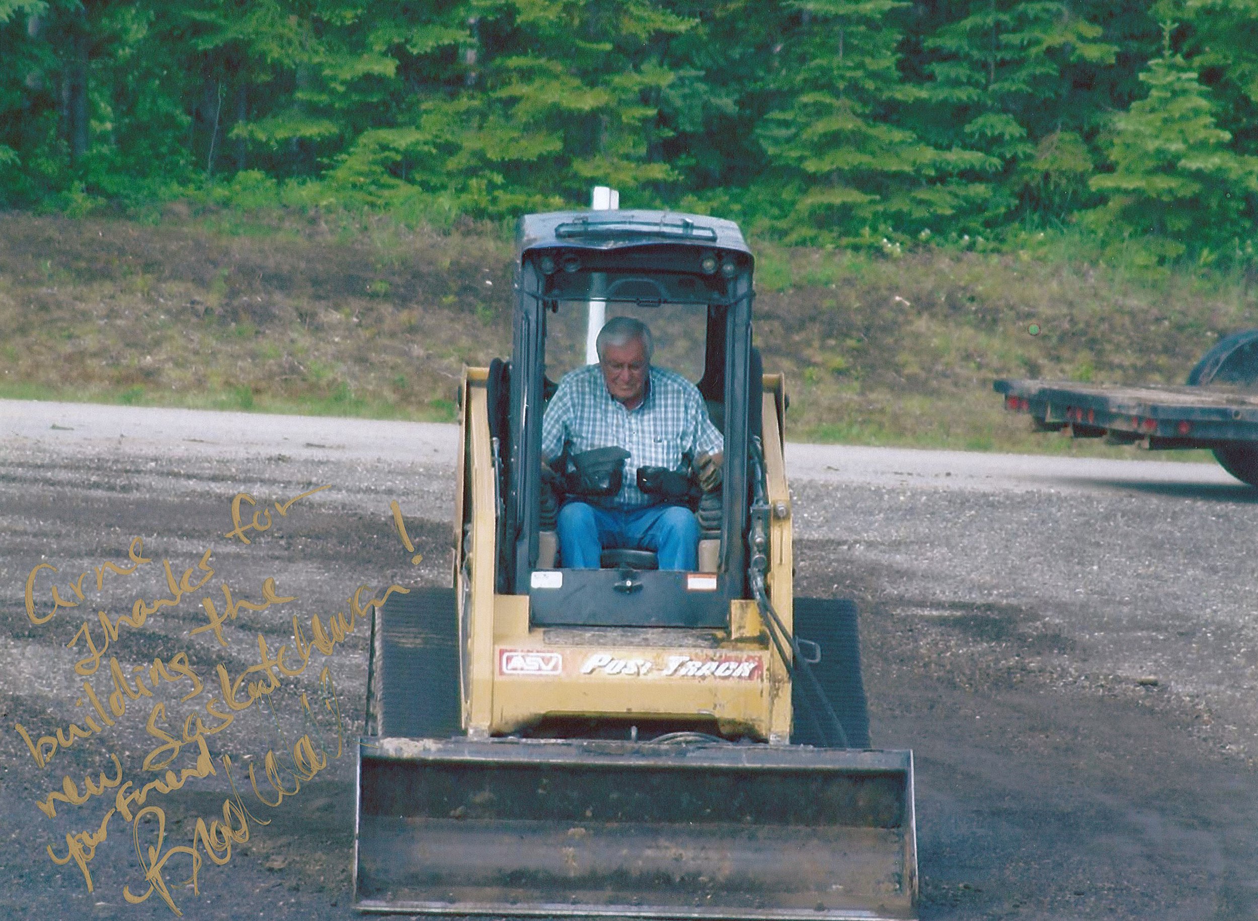 A man operating a small yellow bulldozer on a dirt area, with a background of green trees and a flatbed trailer to the right.