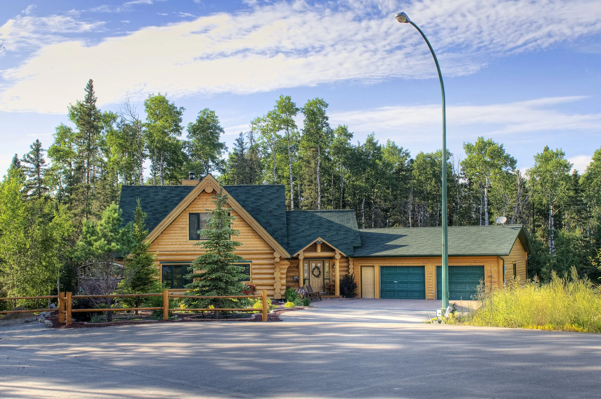 A house with a log cabin style exterior, surrounded by green trees, with a mountain behind and a blue sky with clouds above, a street in front, and a green streetlamp on the right side.