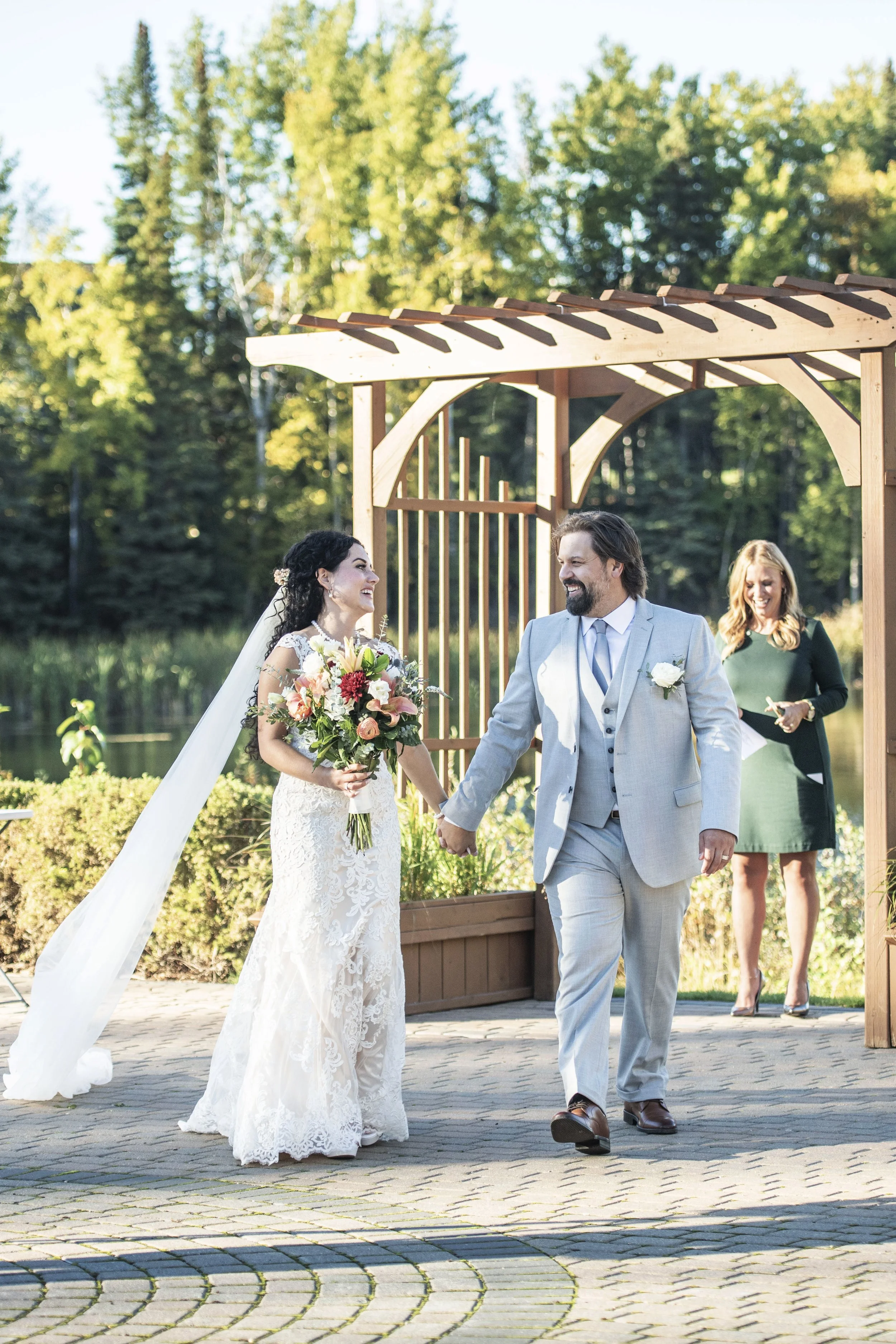 A bride and groom holding hands during their outdoor wedding ceremony, with a woman officiant smiling in the background, under a wooden pergola with green trees and sunlight.