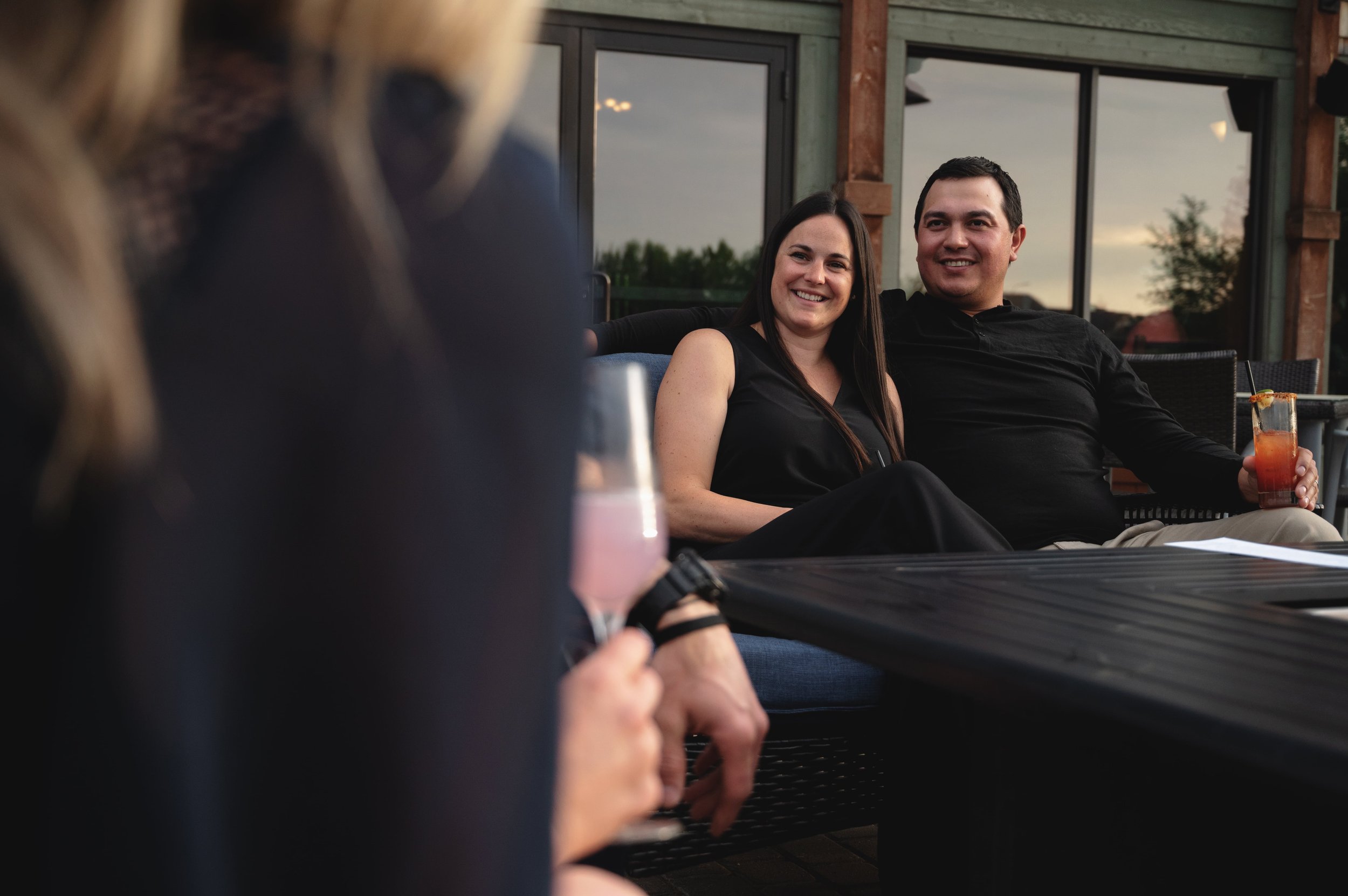 A smiling couple sitting on a patio couch, with the woman wearing a black top and the man wearing a black shirt, holding a drink in a glass with a straw. The background shows large glass windows reflecting the sky and trees at sunset.