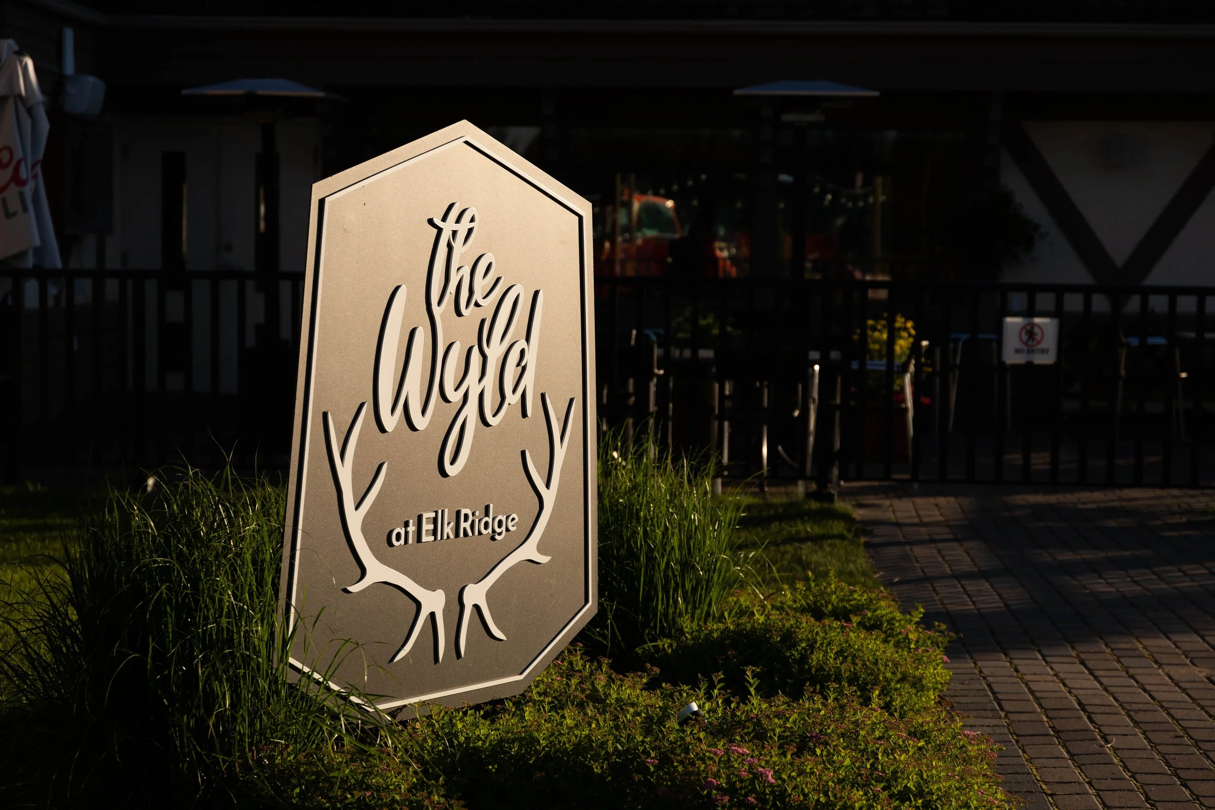Sign reading 'The Lodge at Elk Ridge' with a tree graphic, outside on a lawn with grass and plants, in front of a deck or patio area with umbrellas and a fence.