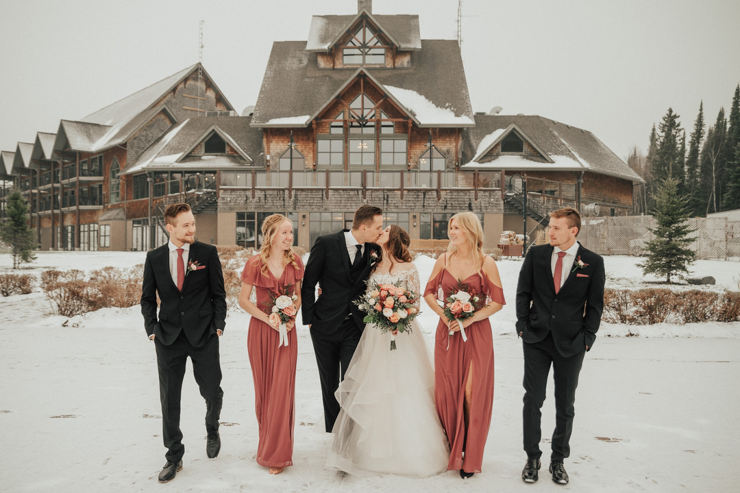 Wedding party standing on snowy ground in front of a large house, with two men in suits and three women in dresses, one of whom is the bride holding a bouquet, kissing a groom.