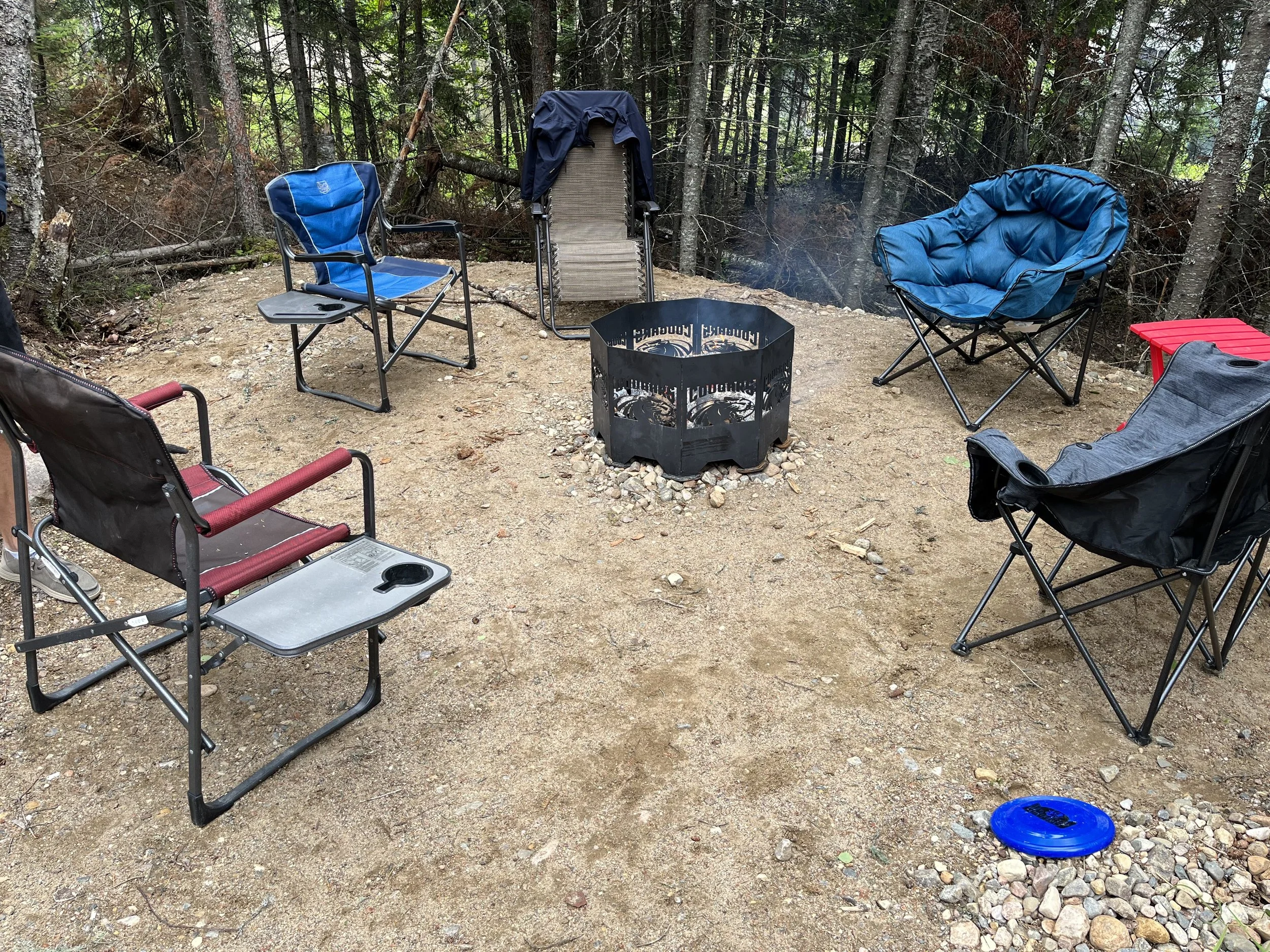 A campsite in a forest with chairs arranged around a fire pit. There are six chairs, including a blue director's chair, a blue padded chair, and a red and black chair. A small red table, a blue frisbee, and a black fire ring are also present. Tall trees surround the area, creating a secluded outdoor setting.