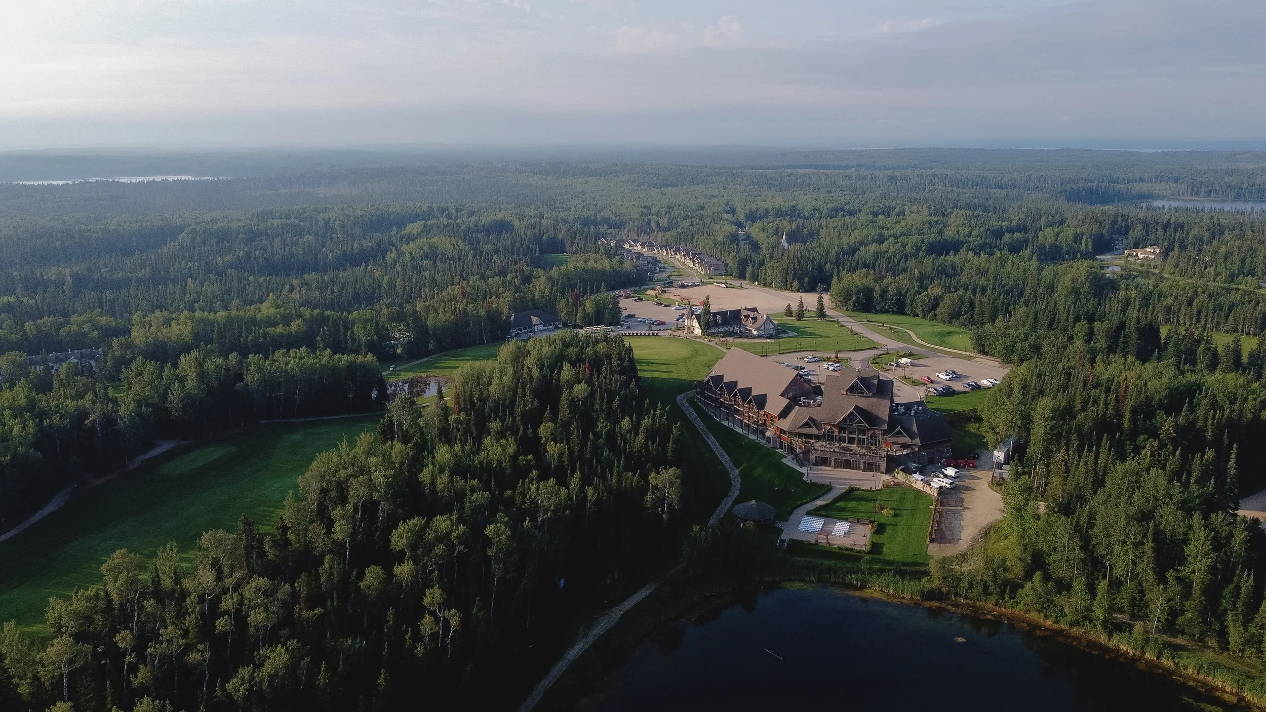 Aerial view of a large resort with multiple buildings, surrounded by dense forest with a lake in the distance, under a cloudy sky.