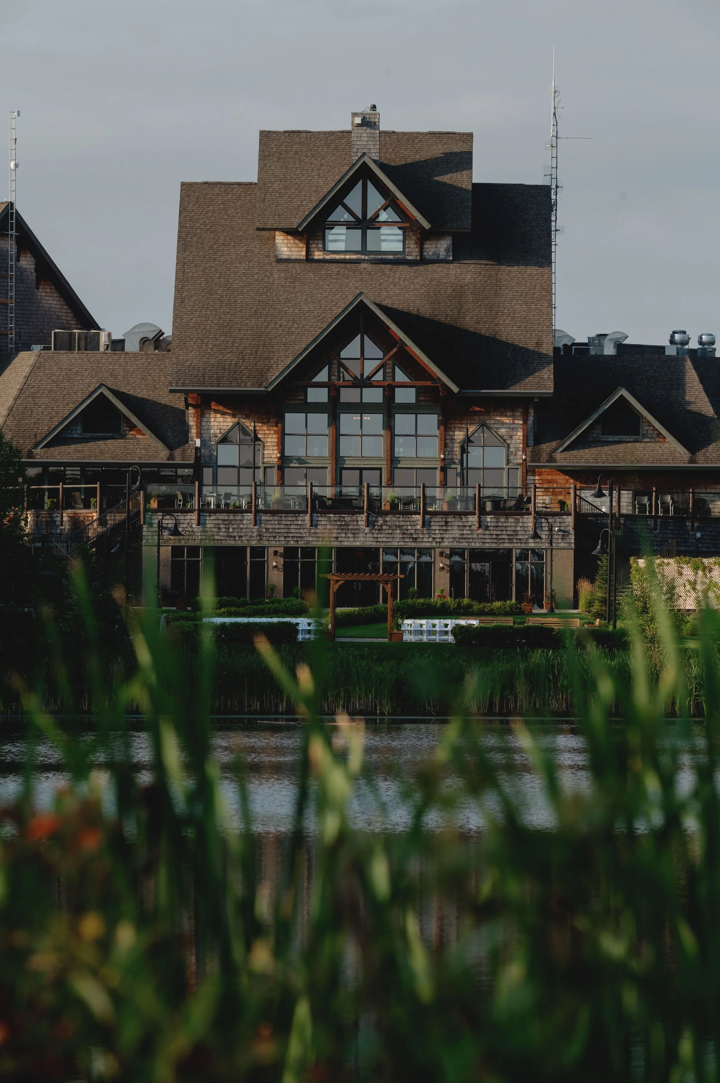 A large residential house with various pitched roofs, a balcony, large windows, and a deck, situated near a body of water with greenery in the foreground.