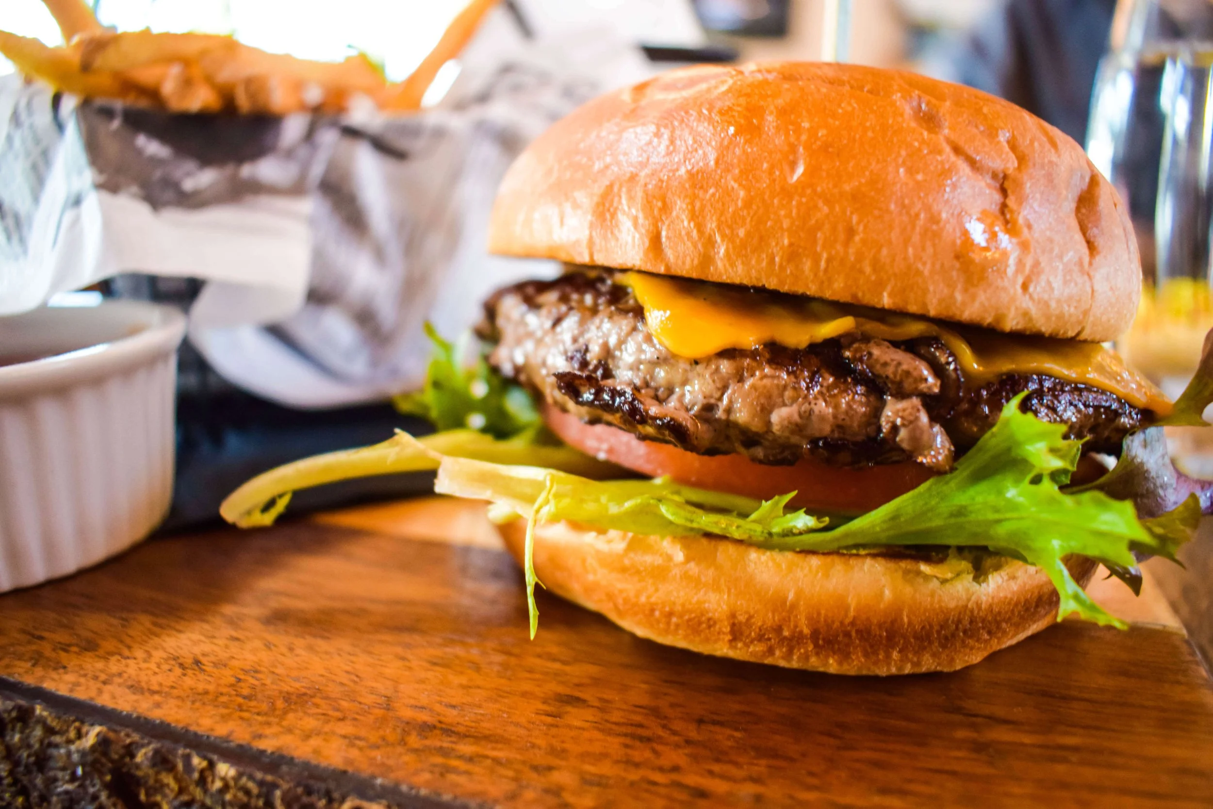 Close-up of a cheeseburger with lettuce, cheese, beef patty, and a bun, on a wooden plate in a restaurant setting.