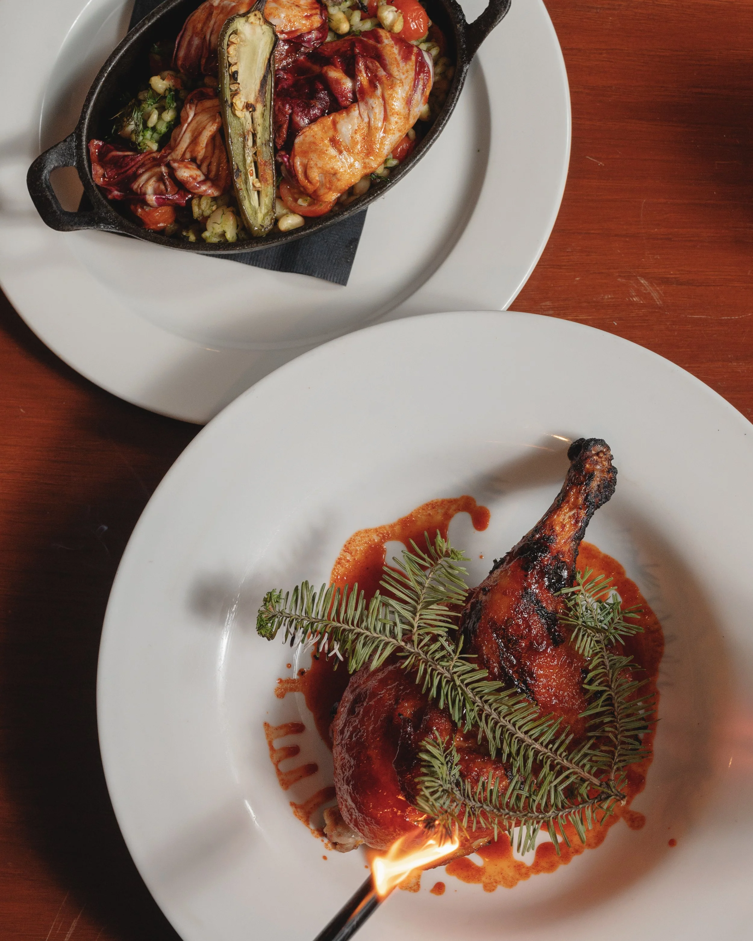 A plate with a cooked chicken leg topped with sauce and garnished with green herbs, placed on a white plate on a wooden table. A flame is visible at the bottom of the image, indicating that the dish may have been flambéed.