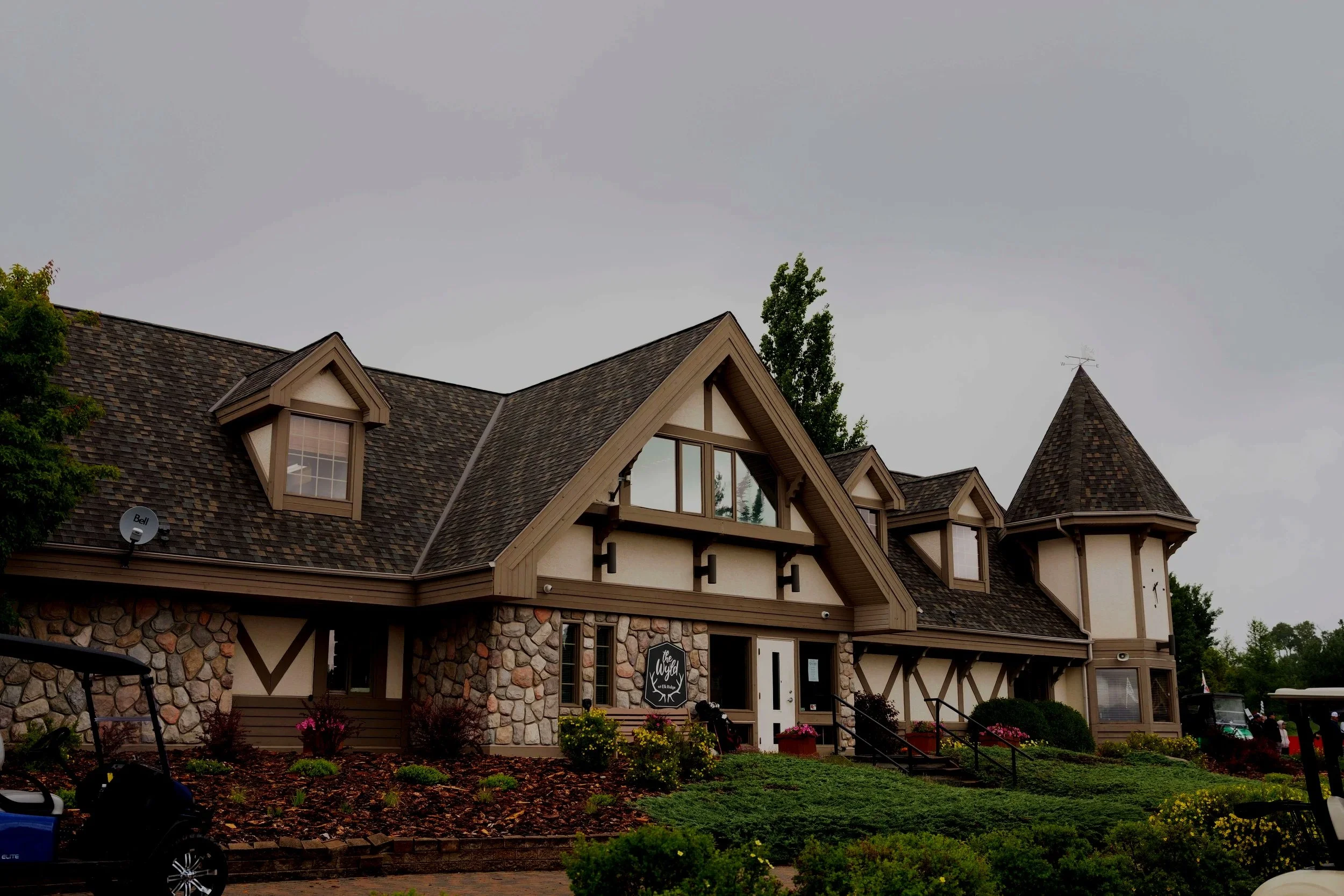 A large house with a stone and wood exterior, steep gable roofs, small dormer windows, and a conical turret. There are trees, flowers, and a clean front yard with a sidewalk.