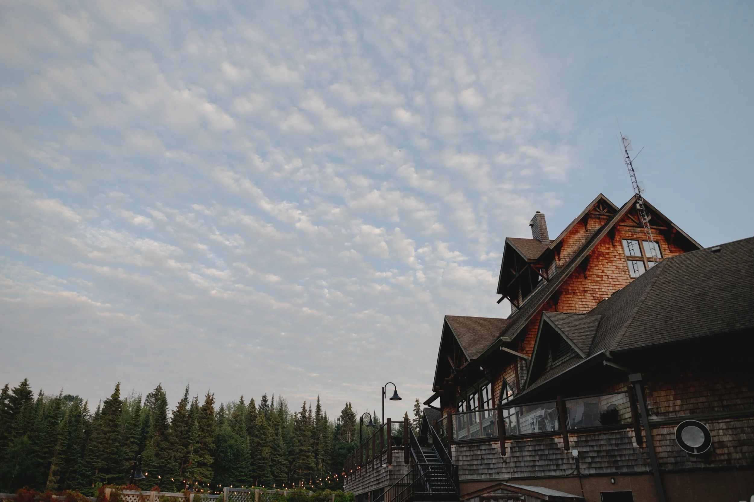 A multi-story house with a steep roof, large windows, and a brick chimney, surrounded by tall trees under a partly cloudy sky.