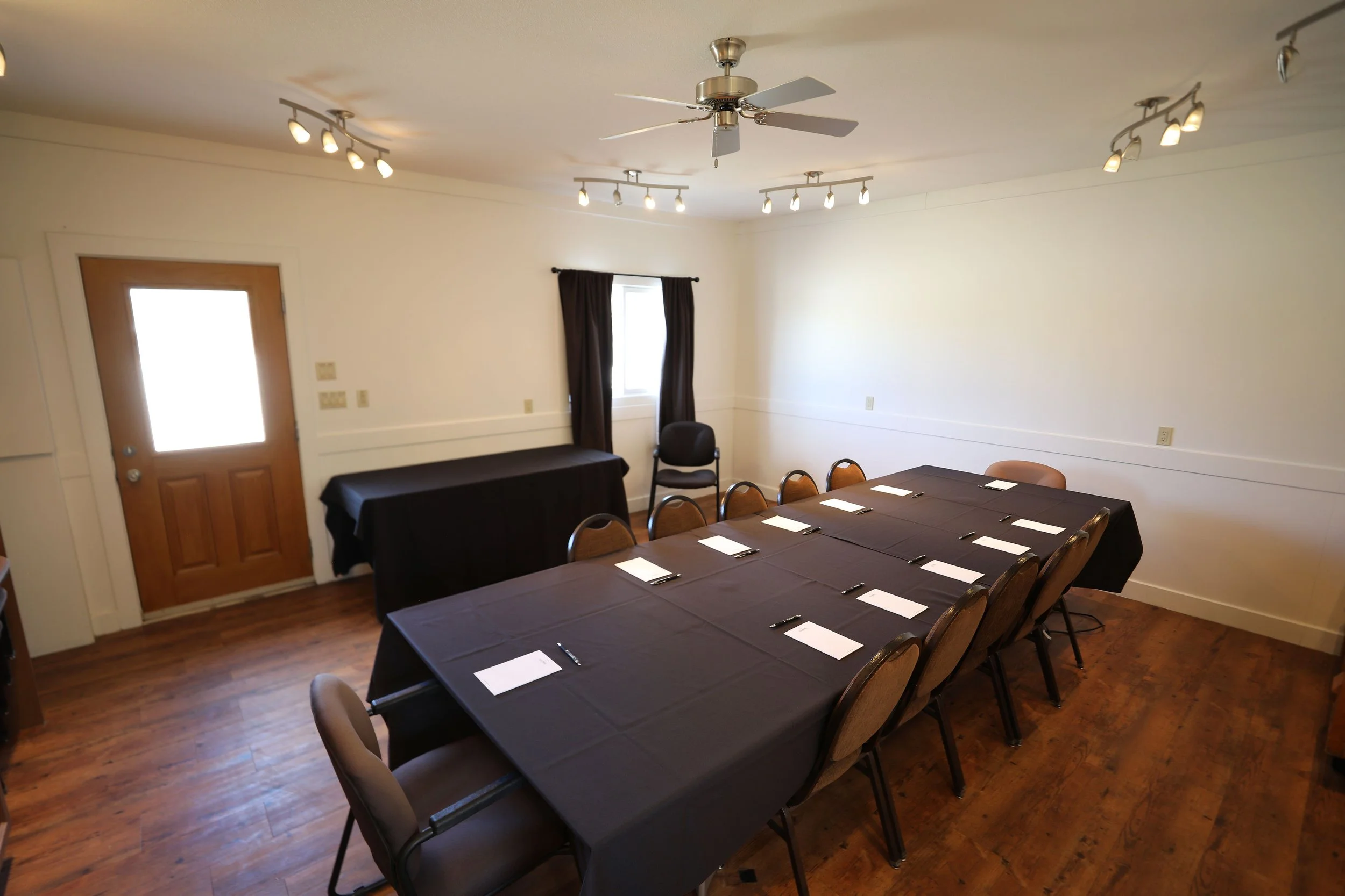 Conference room with a long table covered with a black tablecloth, surrounded by chairs, set for a meeting, with papers and pens. There is a door, a window with curtains, and a white wall. The ceiling has multiple track lights and a ceiling fan.