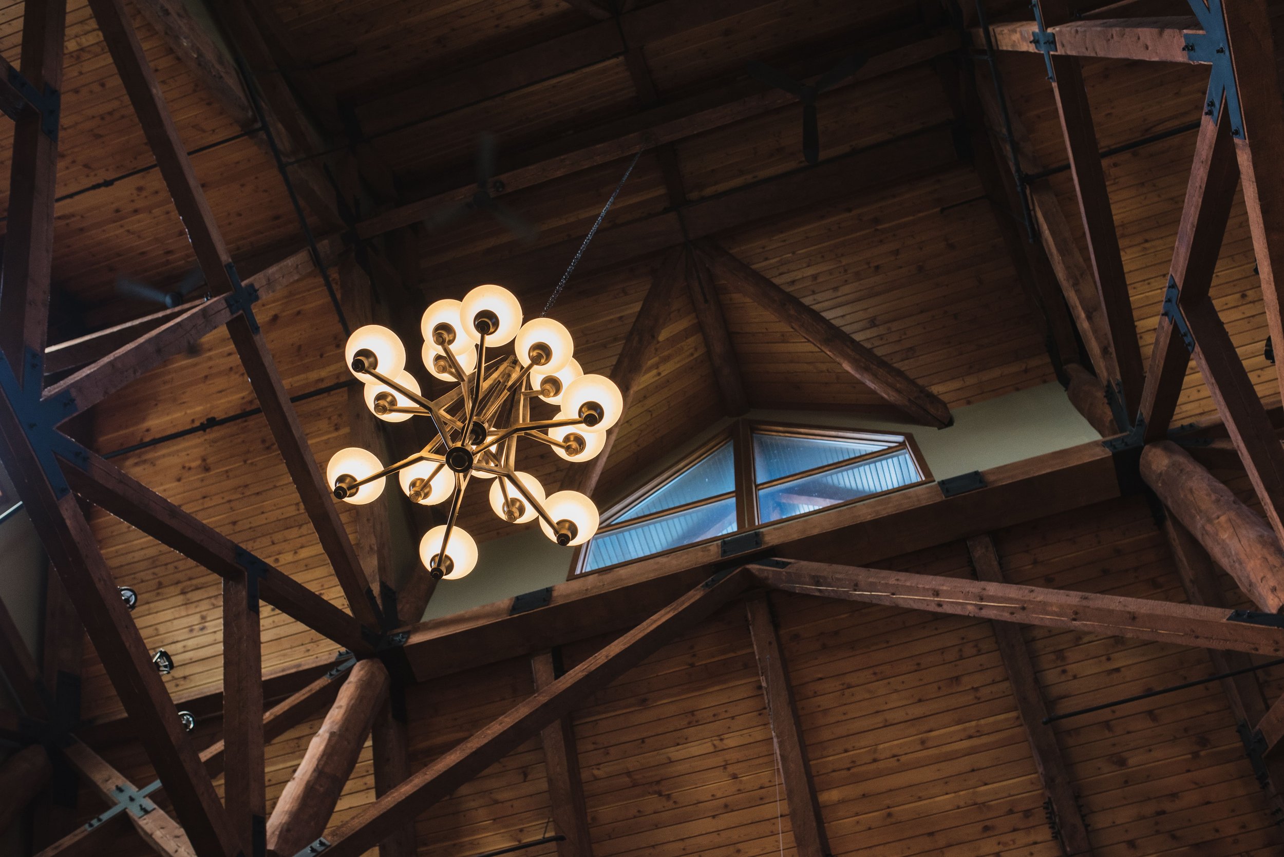 Interior view of a wooden ceiling with a modern chandelier hanging in the center, and a window in the background.