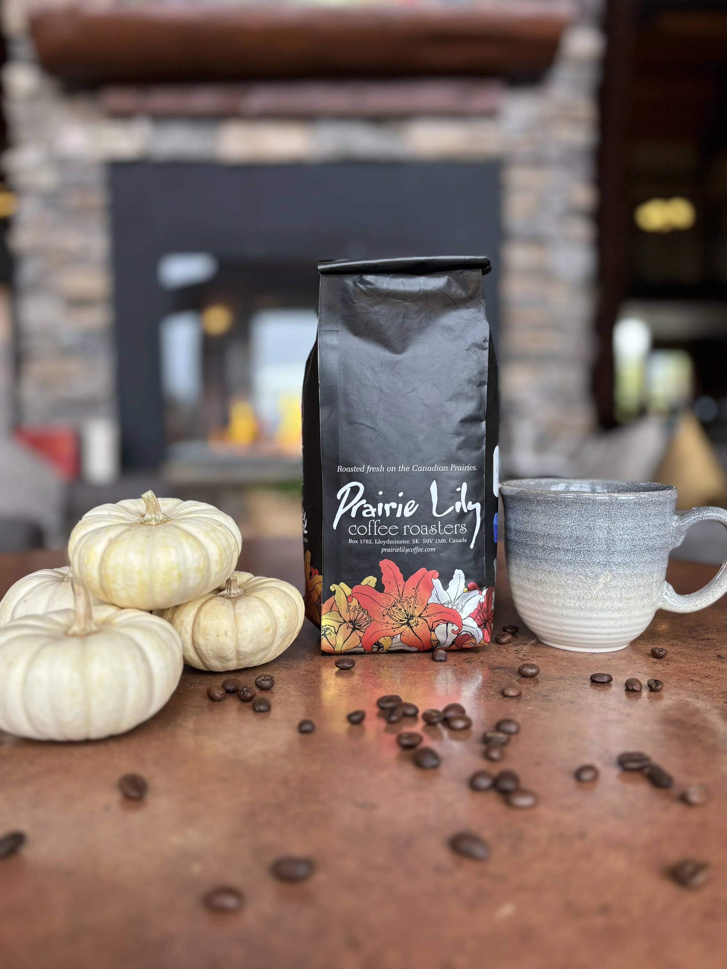A cluster of small white pumpkins, a bag of Prairie Lily coffee, a gray ceramic mug, and scattered coffee beans on a wooden surface, with a blurred fireplace in the background.