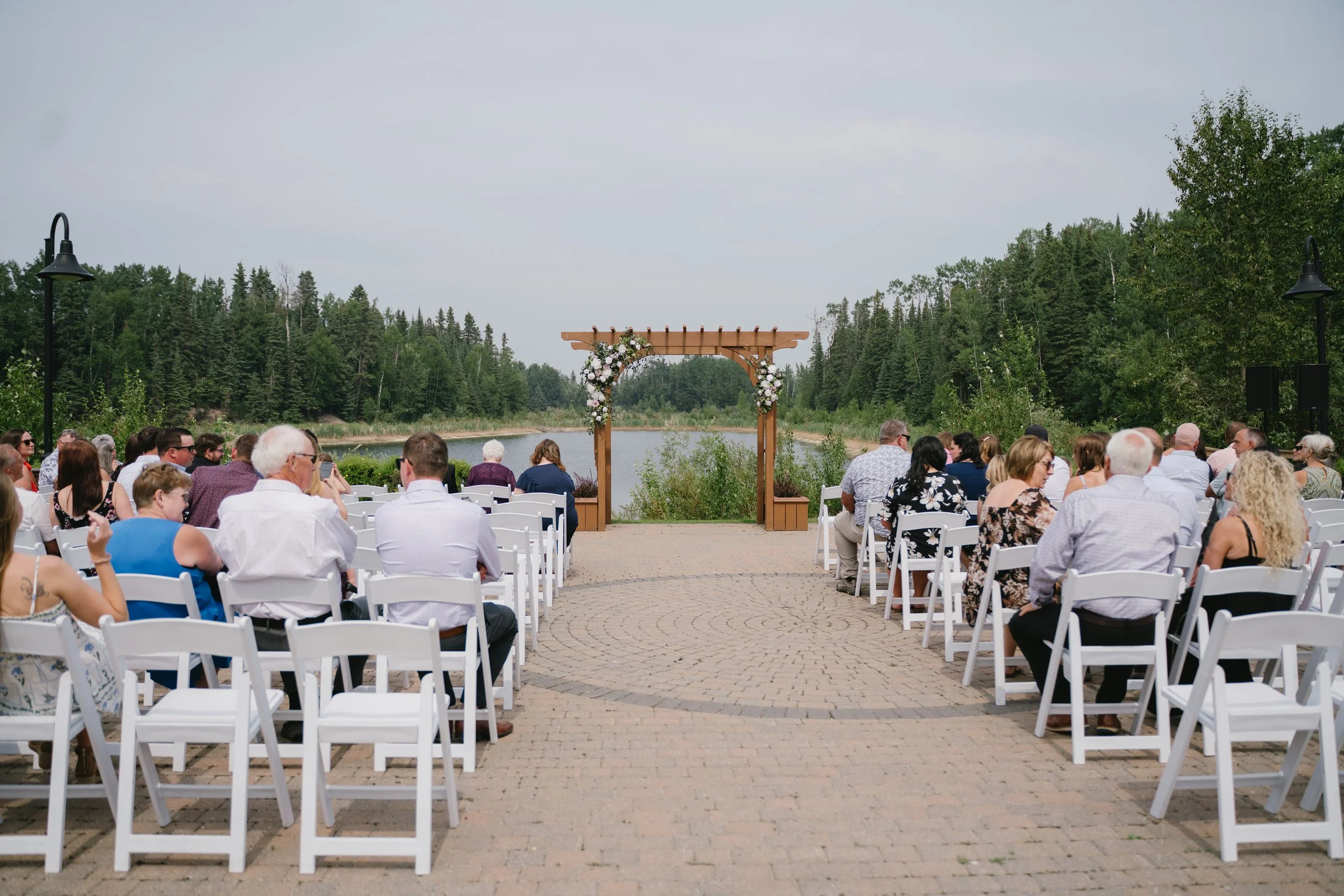 An outdoor wedding ceremony by a lake with a wooden arch decorated with flowers. Guests are seated on white chairs, and there are trees and lampposts along the venue.