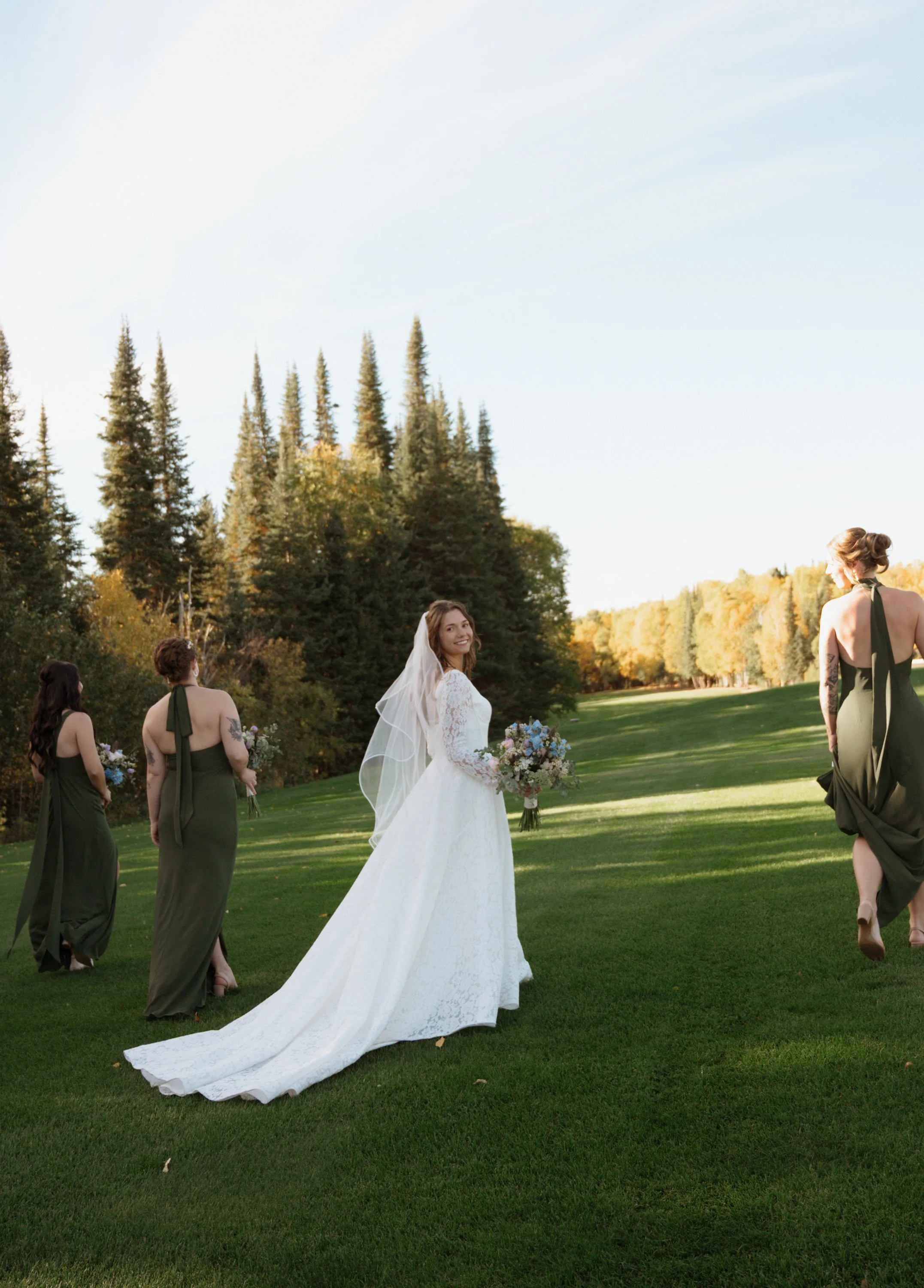 A bride in a white wedding gown stands outdoors on a grassy field, surrounded by bridesmaids in green dresses. The bridesmaids hold bouquets and walk away from the bride. Trees and a clear sky are in the background.