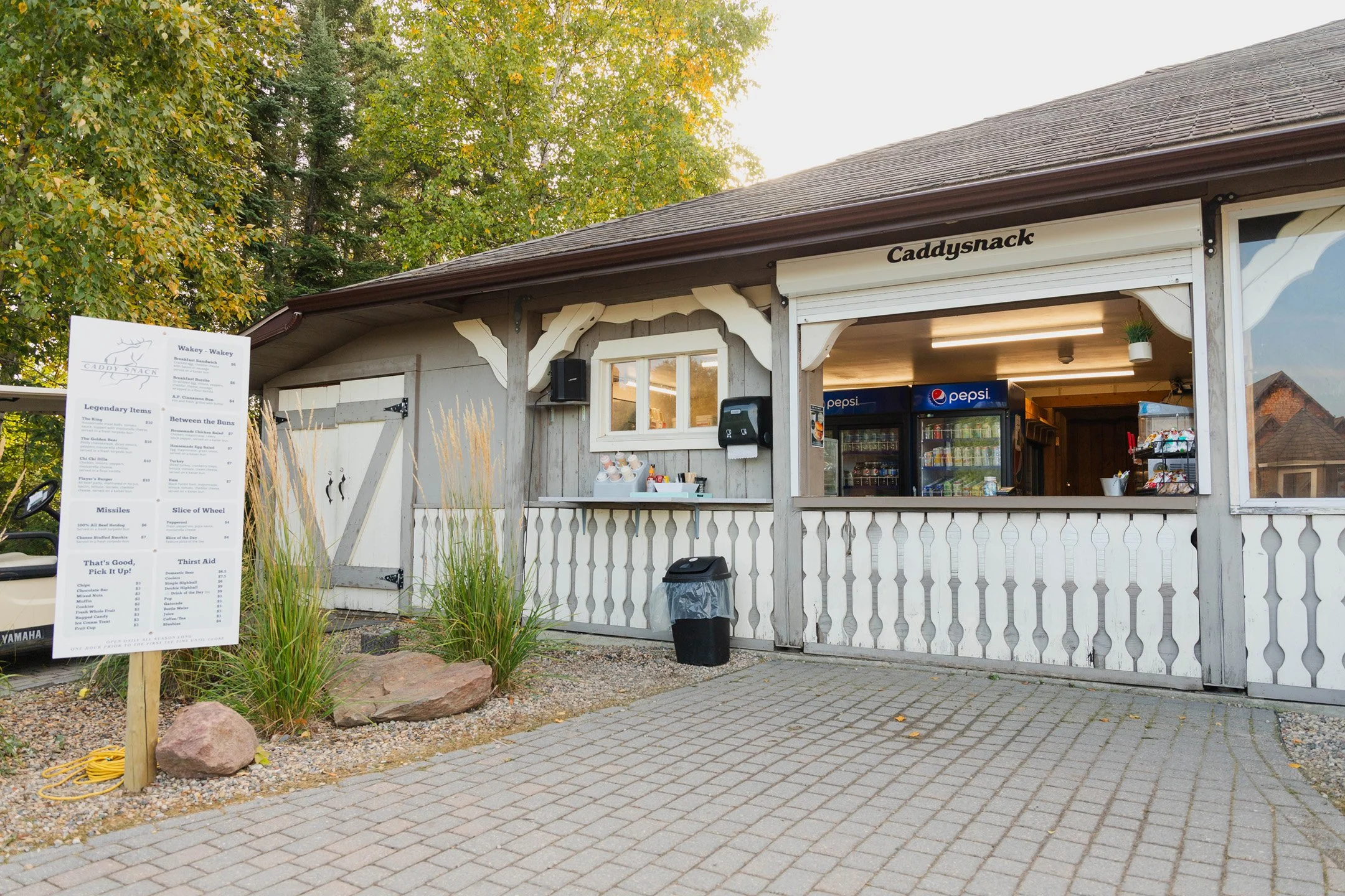 A small wooden snack stand named Caddysnack with a menu board on the left and two Pepsi refrigerators inside. The stand is decorated with white trim and a small plant hanging near the entrance. There is a black trash can in front of the stand, and the area around it is paved with concrete bricks, with trees and a building roof visible in the background.