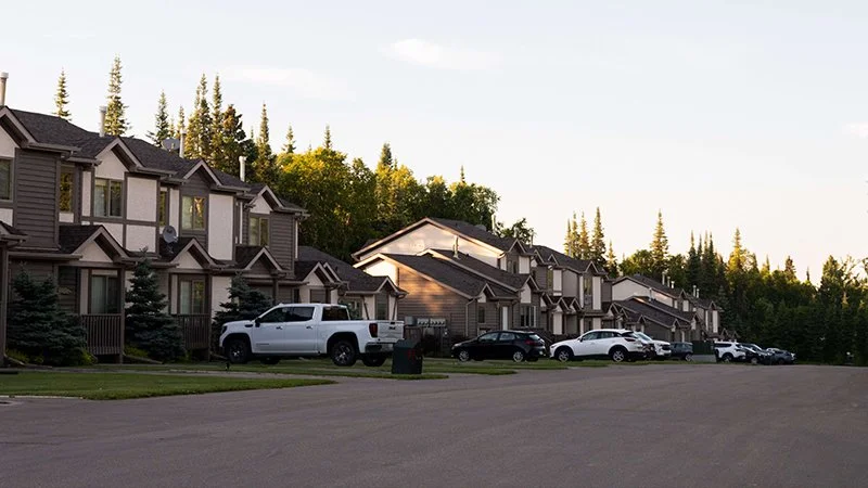 Residential neighborhood with multiple modern houses, parked cars, and a green landscape with trees in the background.