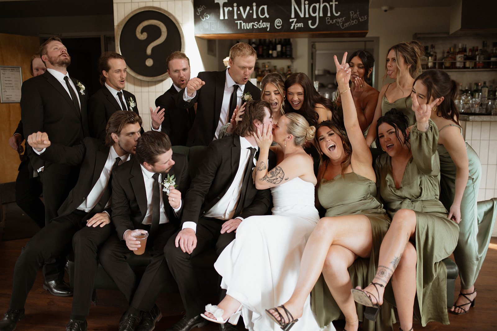 A group of wedding guests and the bride and groom gather around and kiss in a celebratory moment at a wedding reception, some making peace signs and cheering.