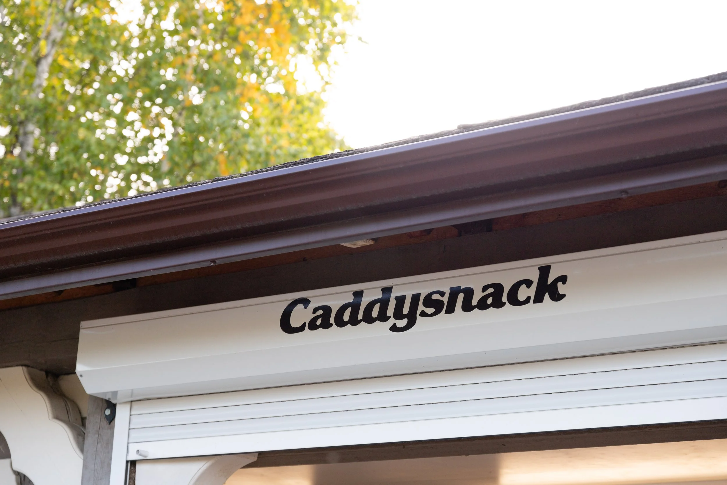 A building with a sign that reads 'Caddysnack' under a roof with trees visible in the background.