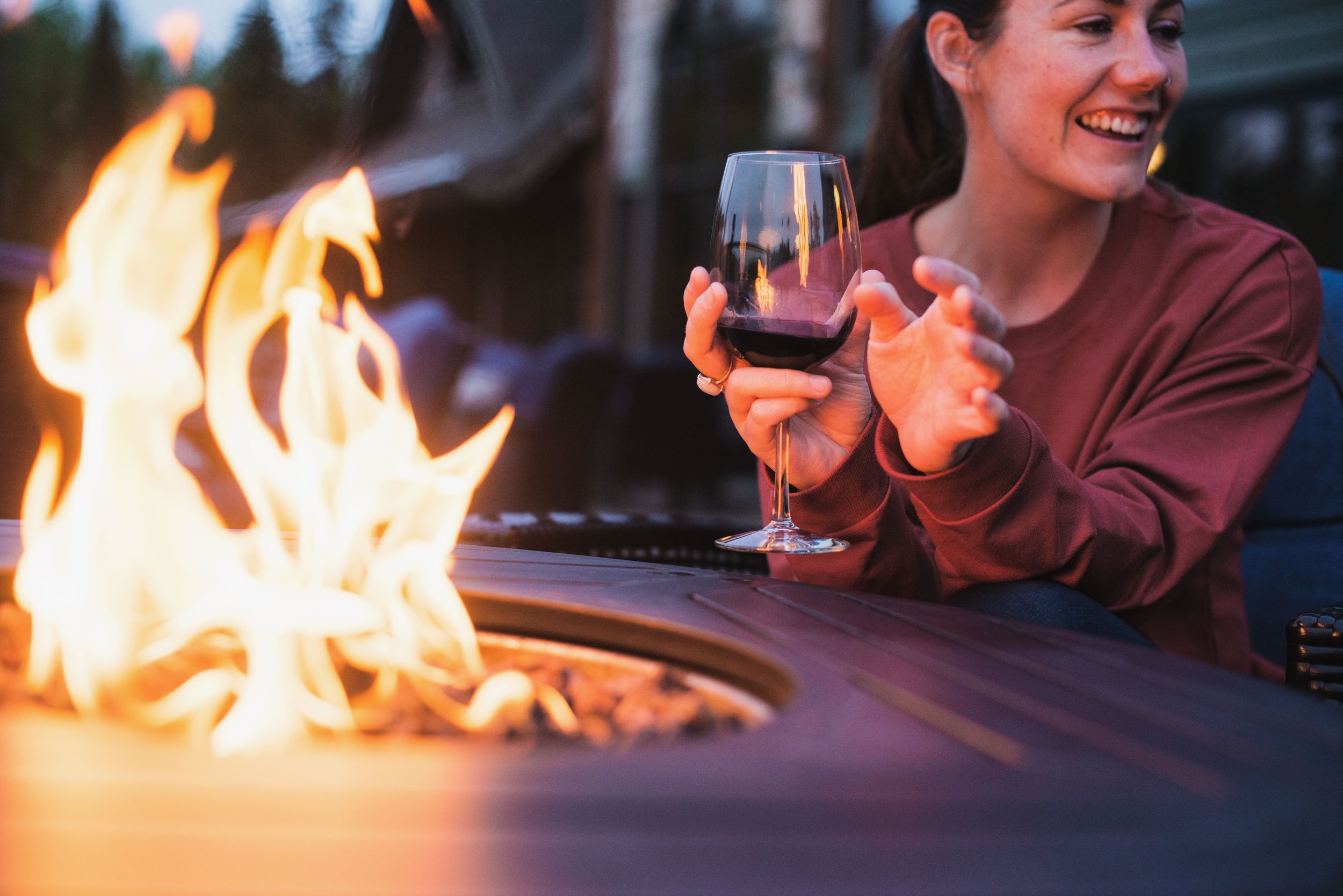 A woman sitting outdoors at a fire pit during dusk, holding a glass of red wine and smiling.