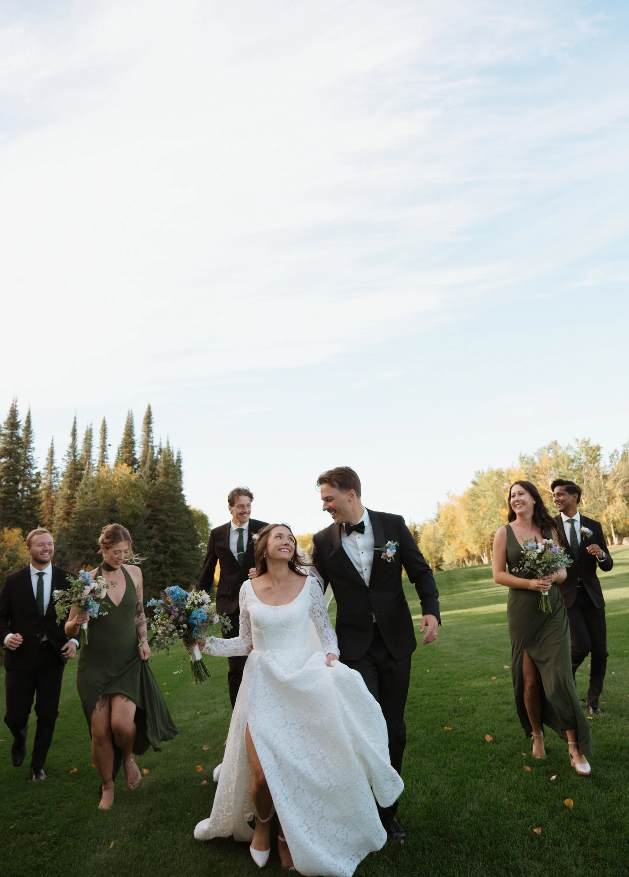 A wedding party, including the bride in a white dress and groom in a black tuxedo, walking on green grass outdoors surrounded by bridesmaids and groomsmen, with trees and a partly cloudy sky in the background.