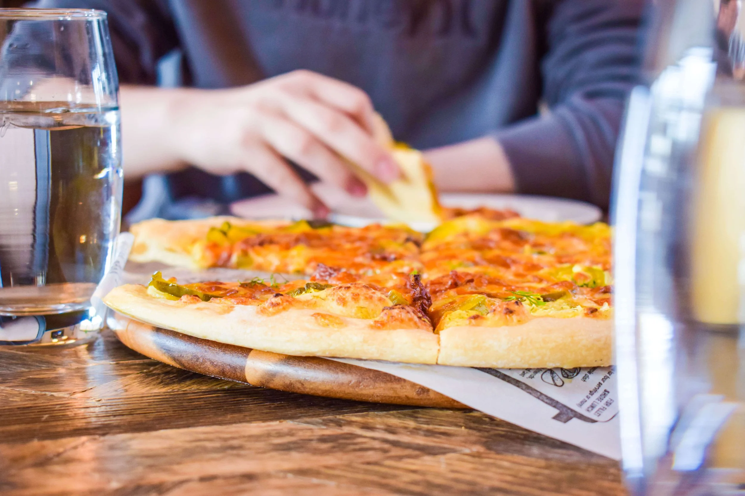 A person in a long-sleeve gray shirt sitting at a table with a fresh pizza topped with green peppers and cheese, and a glass of water.