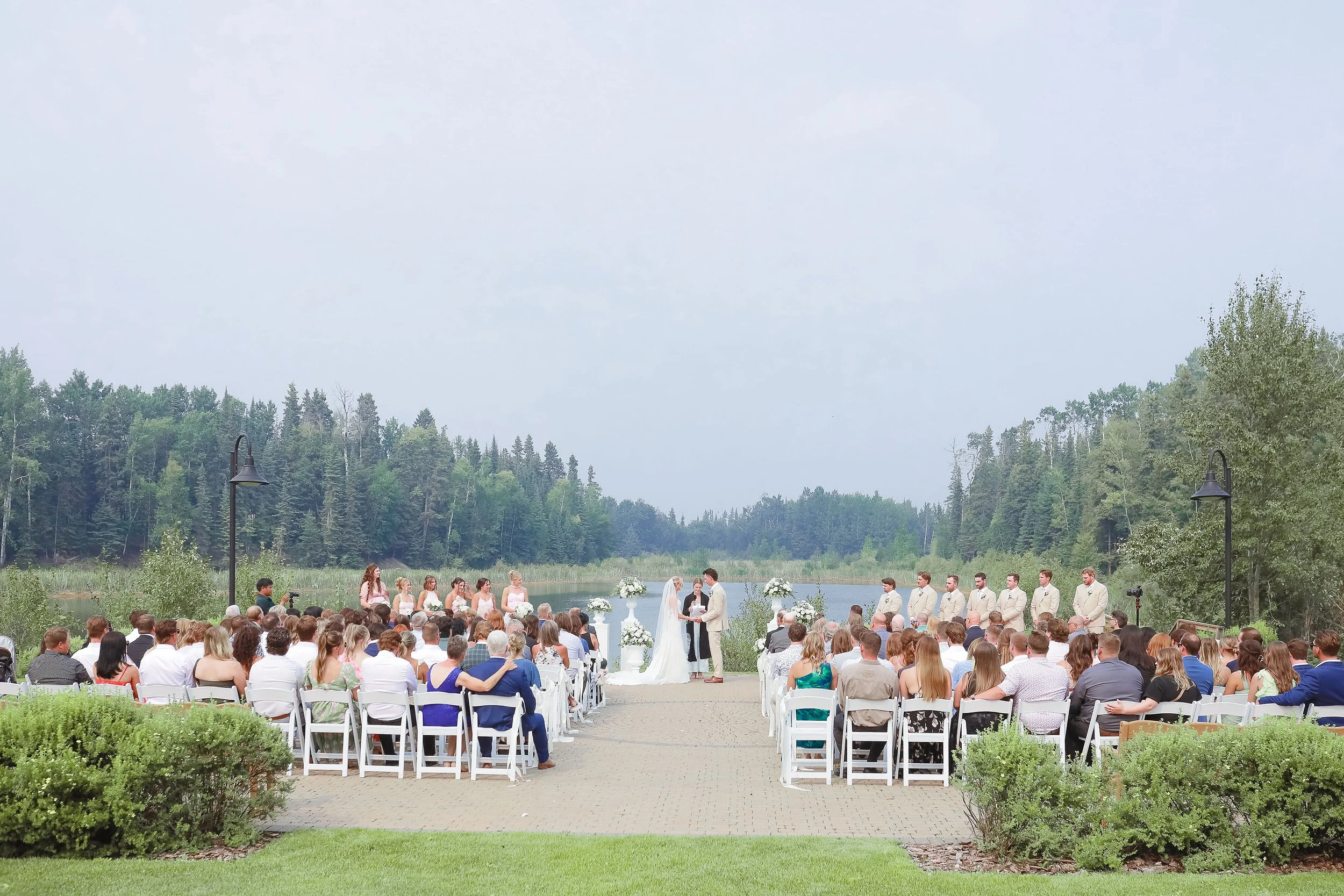 An outdoor wedding ceremony by a river with a backdrop of trees and a cloudy sky. The bride and groom are standing at the altar, surrounded by bridesmaids and groomsmen, while guests sit in white chairs facing the couple.