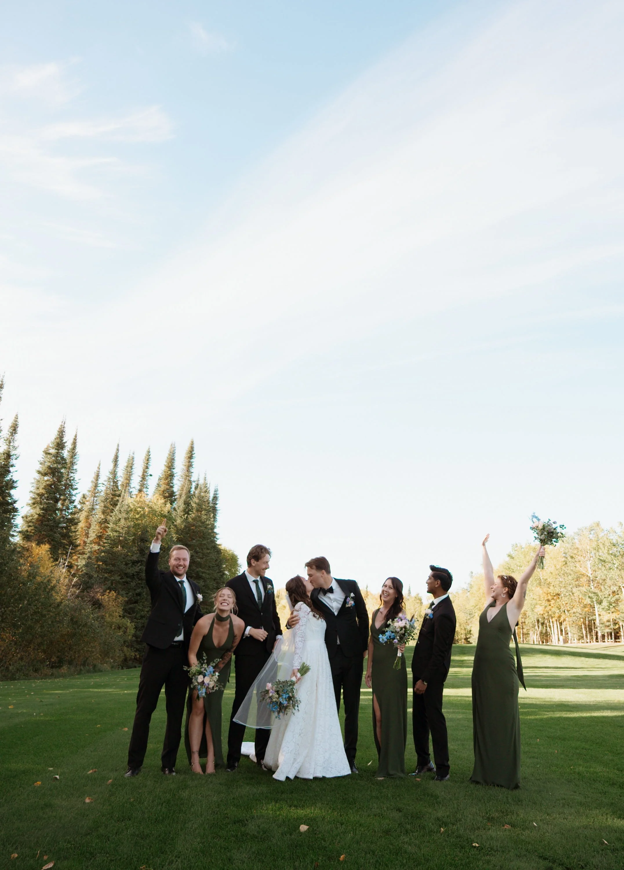 A group of wedding guests celebrating outdoors on a grassy field, with a bride and groom at the center. The groom is wearing a tuxedo and the bride is in a white wedding dress. The guests are dressed formally and are smiling, laughing, and holding flower bouquets, with some raising their hands in celebration. There are tall trees and a clear blue sky in the background.