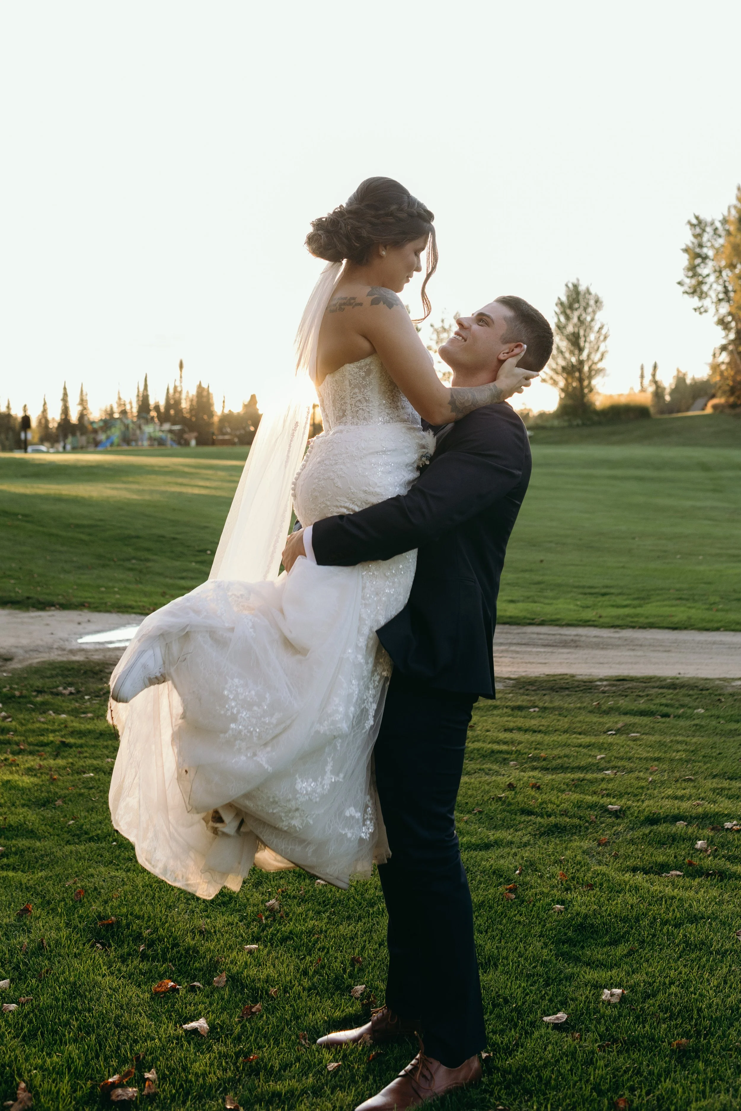 A groom in a black suit and brown shoes lifts a bride in a white lace wedding dress with a long train, holding her in his arms during sunset in a park with green grass and trees in the background.
