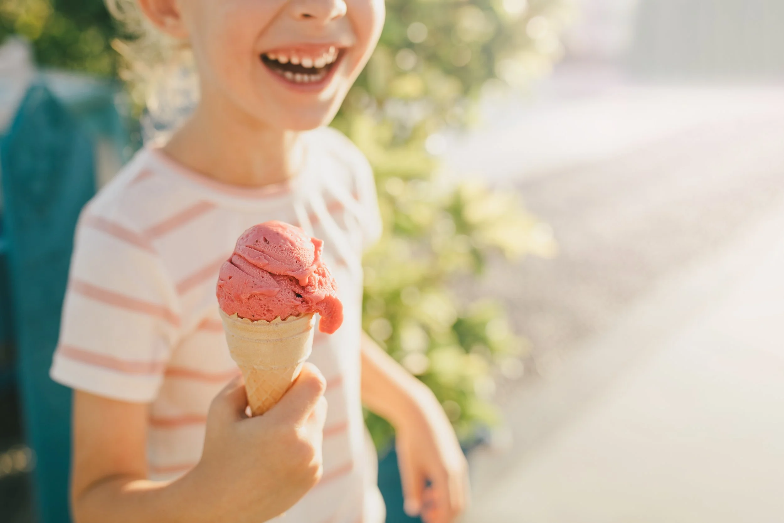 A child smiling and holding a cone of pink ice cream outdoors during daytime, wearing a white and pink striped shirt.