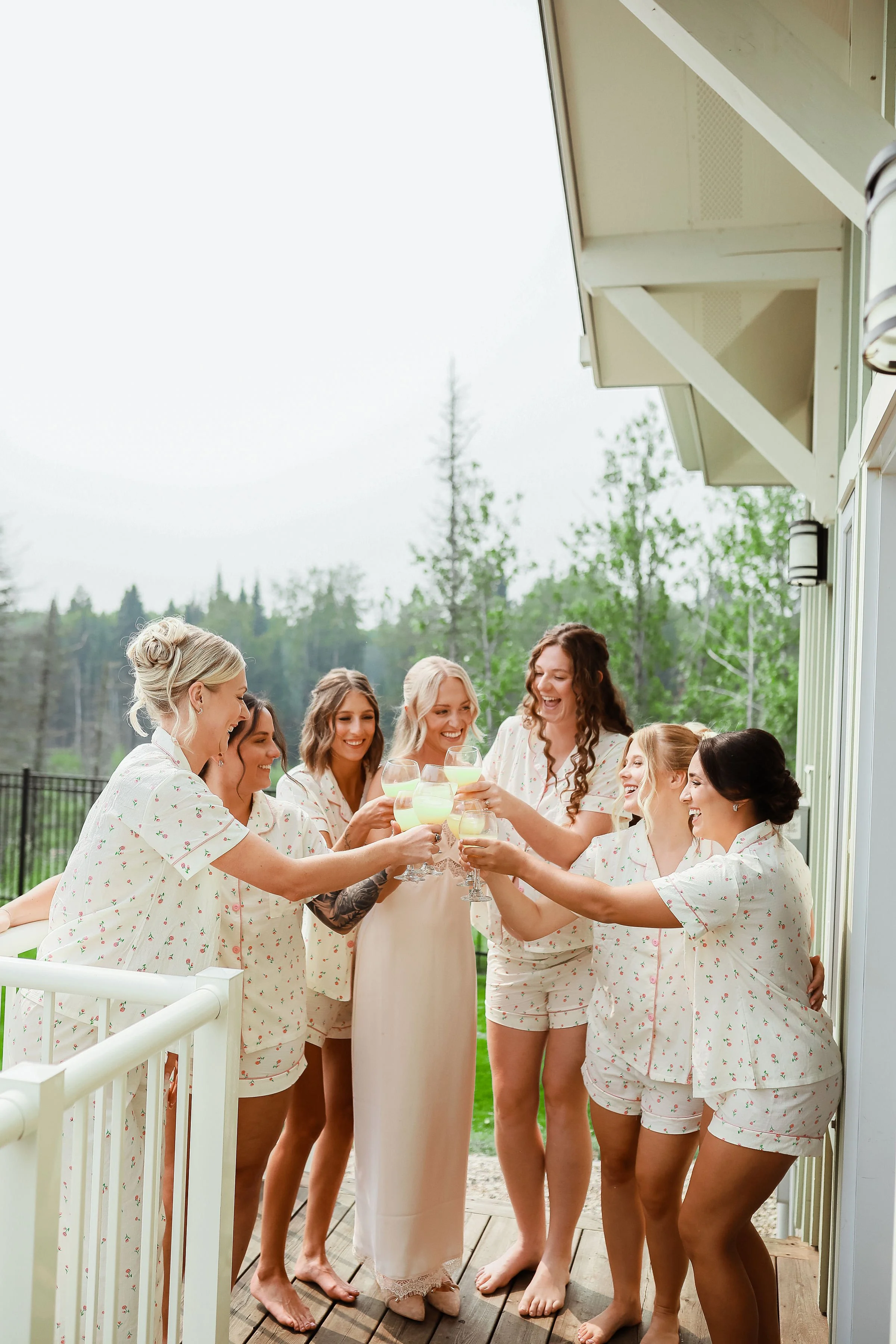A group of women in pajamas are standing outside on a deck, smiling and holding glasses of margaritas in a toast. They are celebrating a special occasion, possibly a bridal shower or similar event, with a background of trees and a cloudy sky.