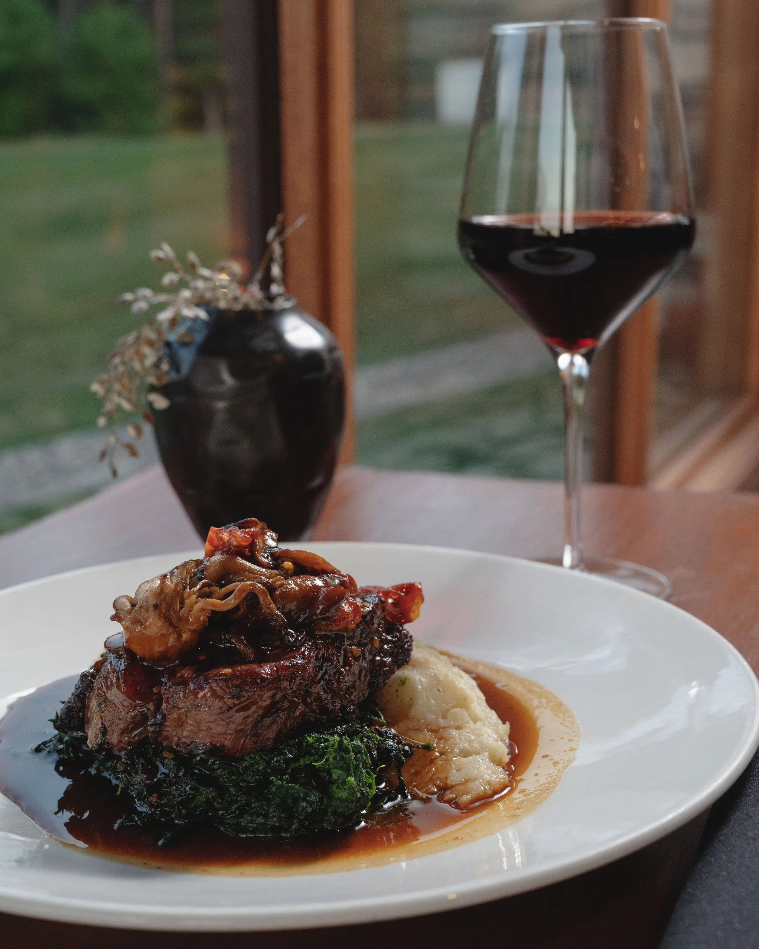A plated serving of meat with a rich sauce, mashed potatoes, and cooked greens, with a glass of red wine next to it, set in front of a window showing greenery outside.