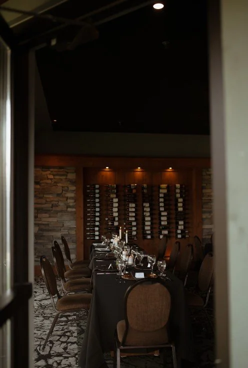 A long dining table set with glasses, plates, and folded napkins, chairs surrounding it, inside a cozy restaurant or wine cellar with stone walls and wine racks in the background.
