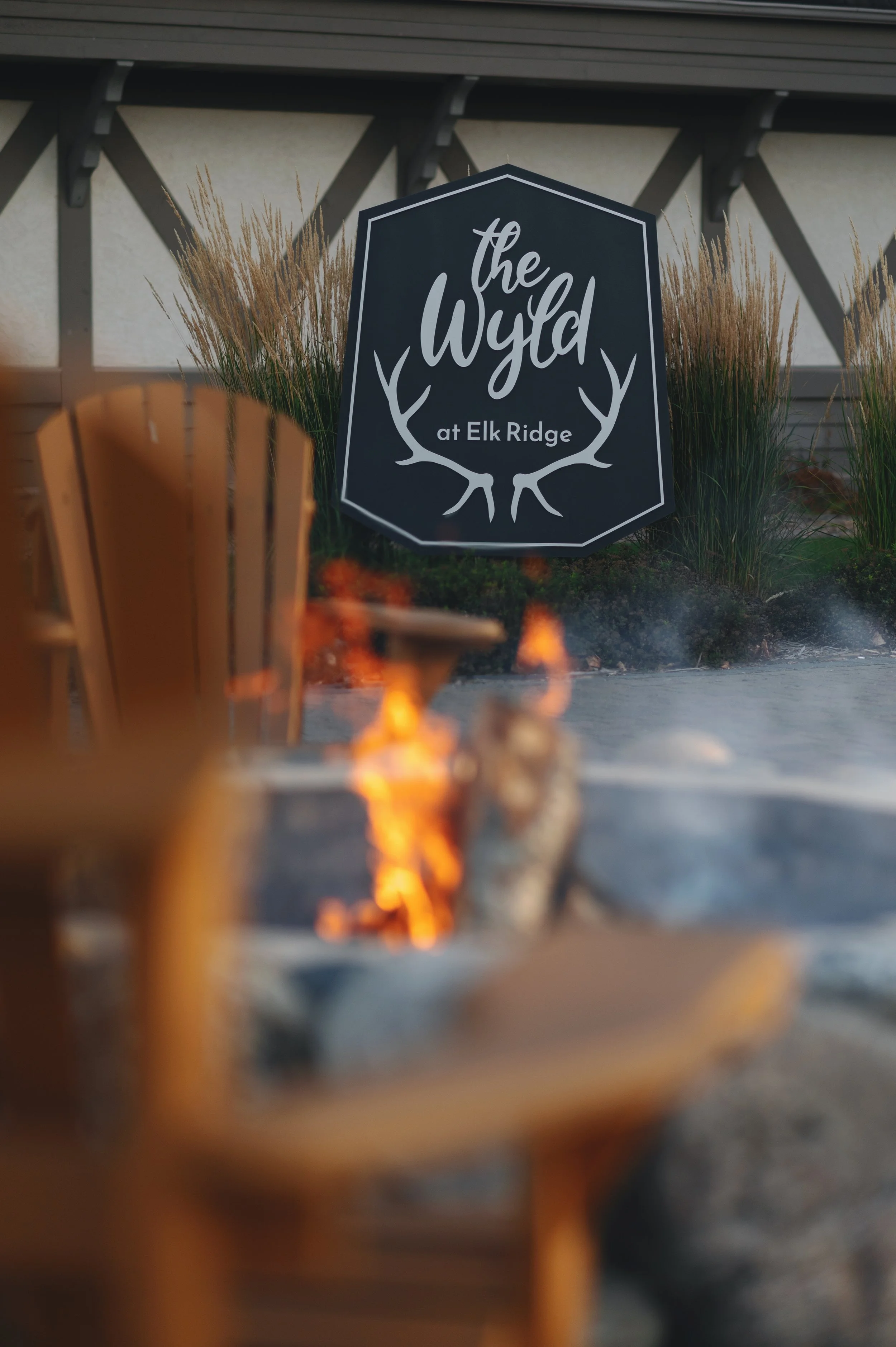 A sign reading 'the Wyo at Elk Ridge' with antler graphics, surrounded by ornamental grasses, with a fire pit in the foreground and part of a building in the background.