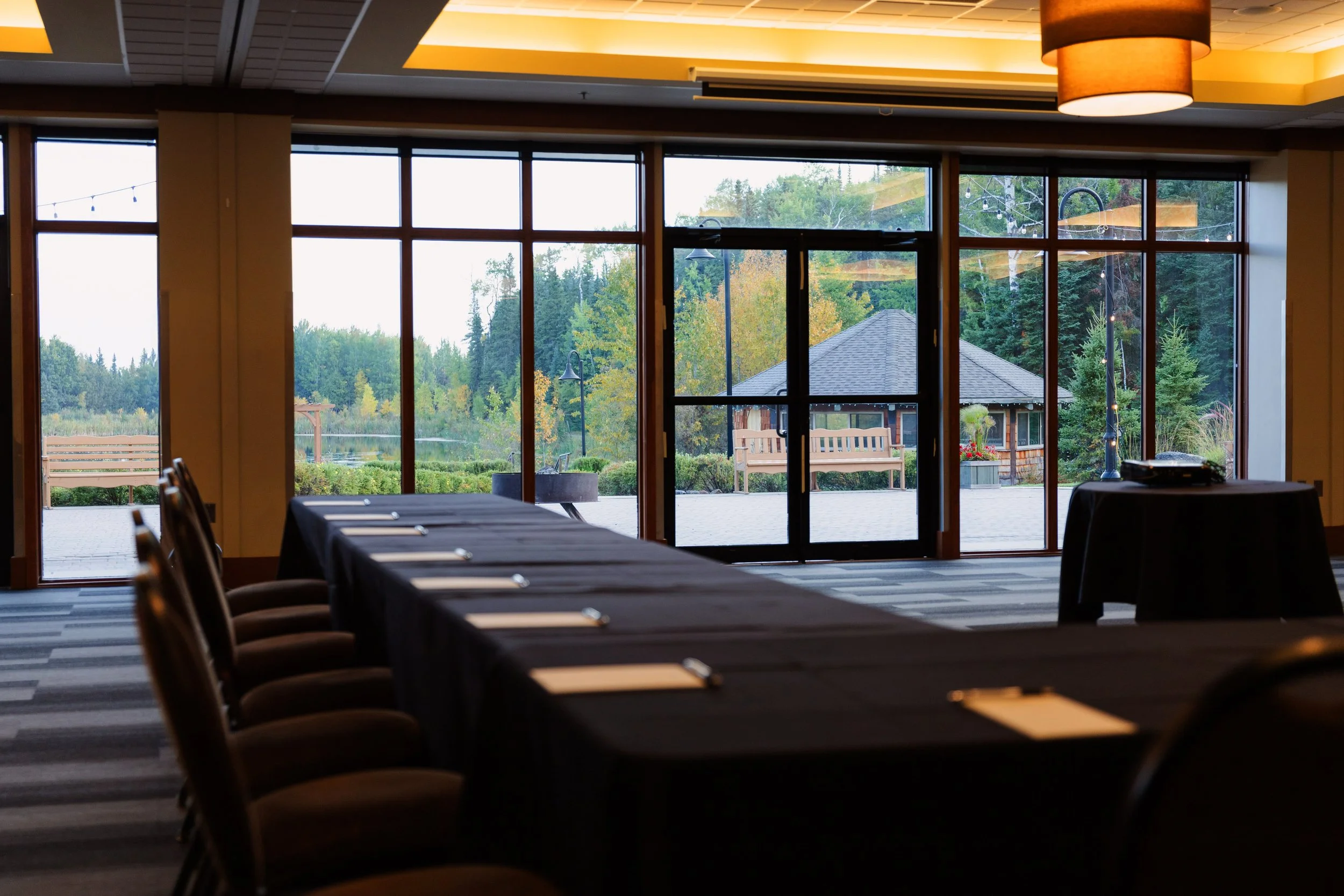 Inside a room with large windows showing an outdoor landscape of trees, a small building, and string lights. There are tables and chairs arranged for a gathering.