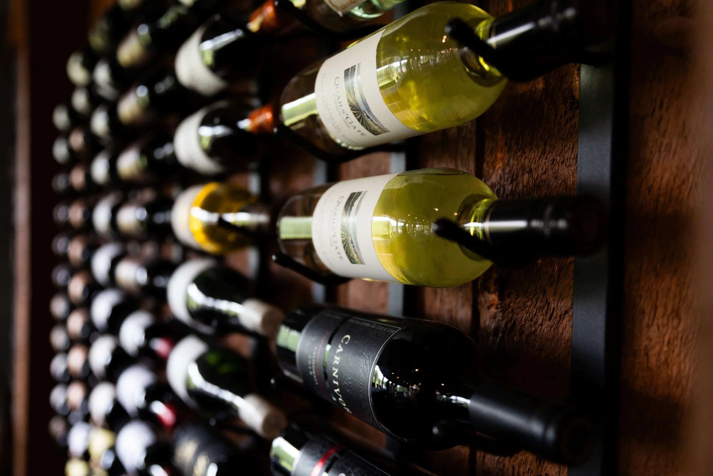 Rows of wine bottles stored horizontally on a rack, with a focus on a few bottles with labels visible, including a bottle with a yellowish wine.