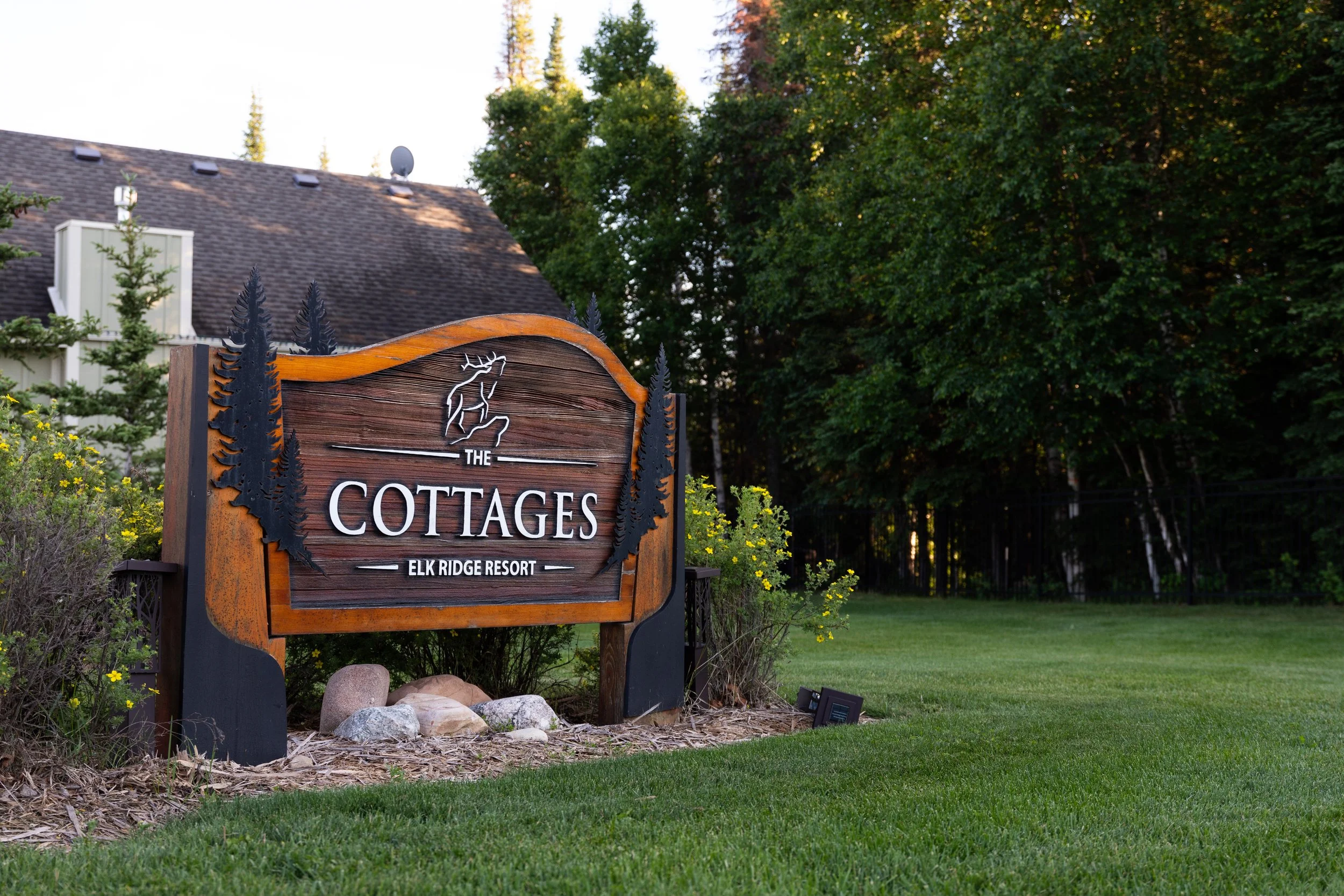 Wooden sign that reads "The Cottages Elk Ridge Resort" with trees and a house in the background.