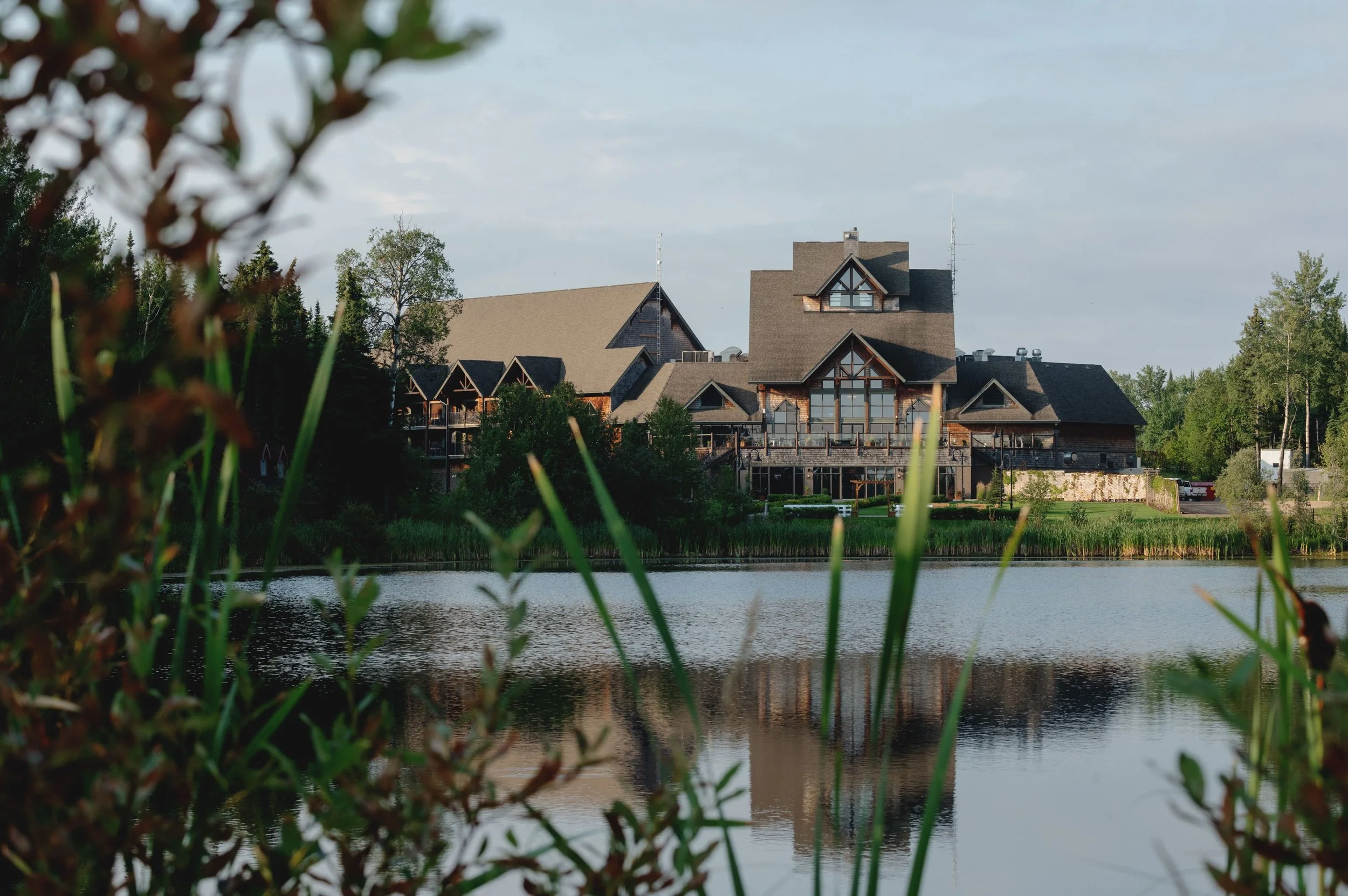 A large lodge-style building with multiple gabled roofs, situated by a body of water, surrounded by trees, with a partly cloudy sky in the background.