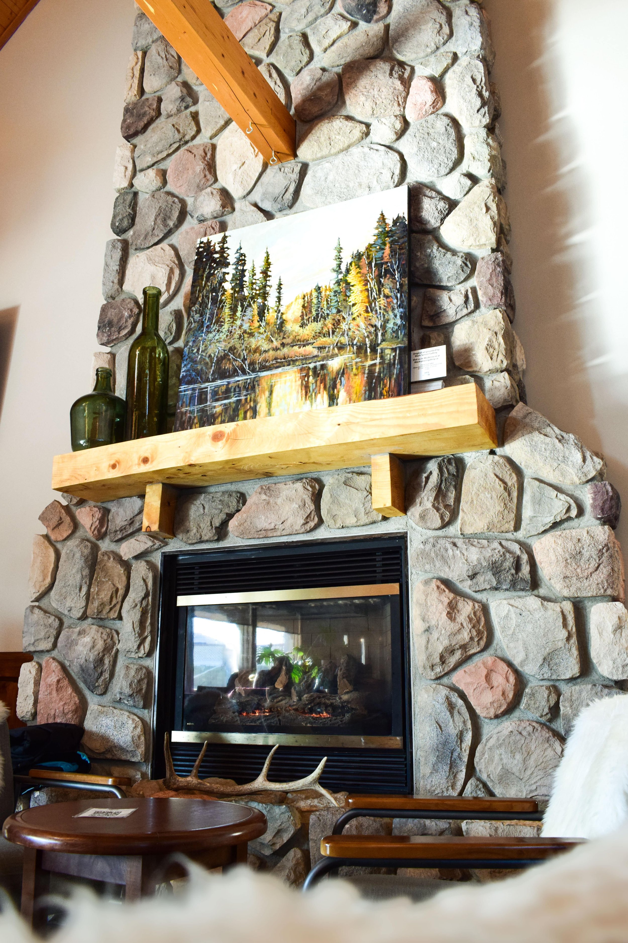 Indoor scene with a stone fireplace and mantle. On the mantle, there are two green glass bottles and a colorful landscape painting of a forest with trees and a river, reflecting sunset or sunrise. Below the mantle, a table with a deer antler on it is visible, along with chairs.