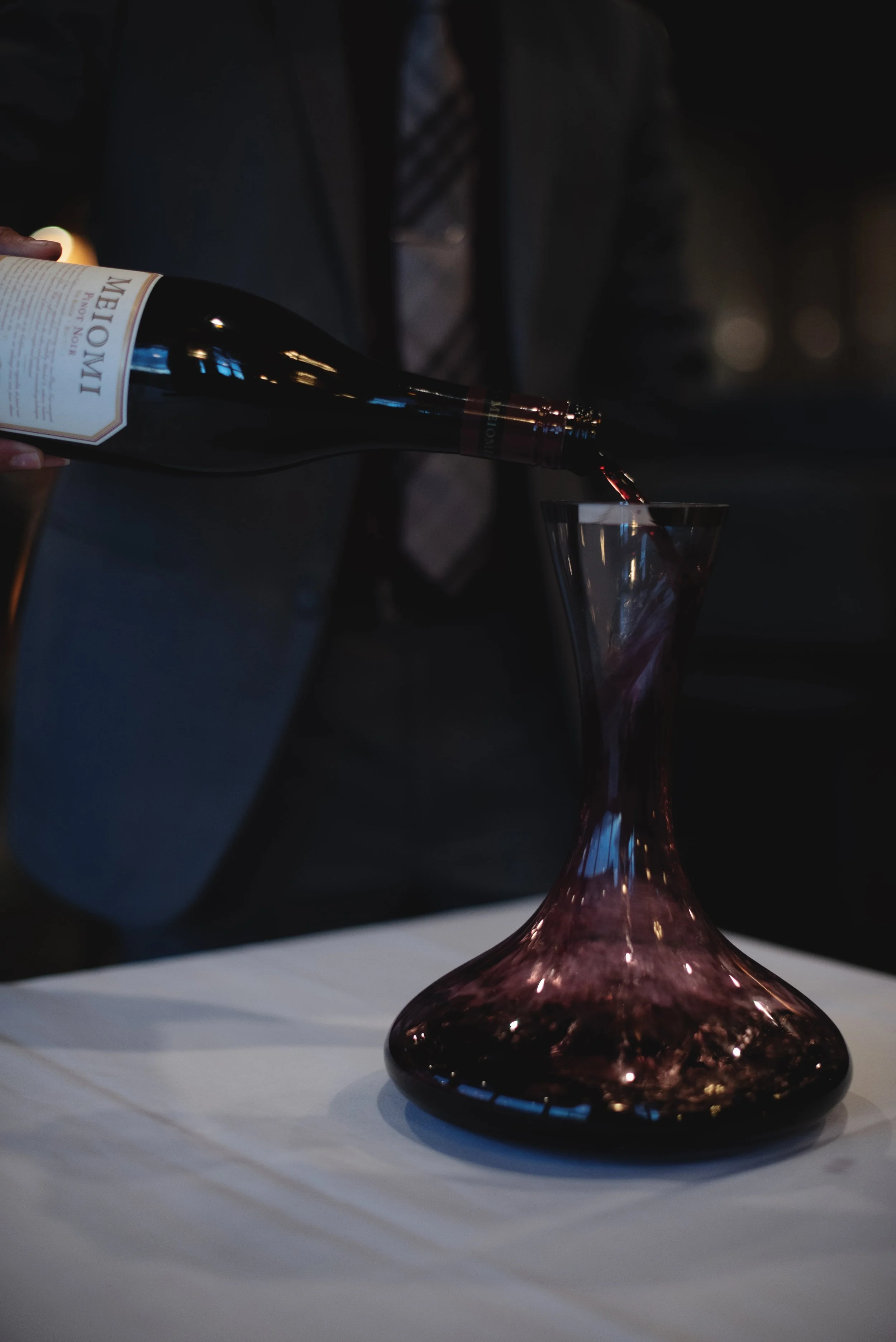 A person is pouring red wine from a bottle into a pink and purple glass on a white surface, with a blurred background of a person in a suit.