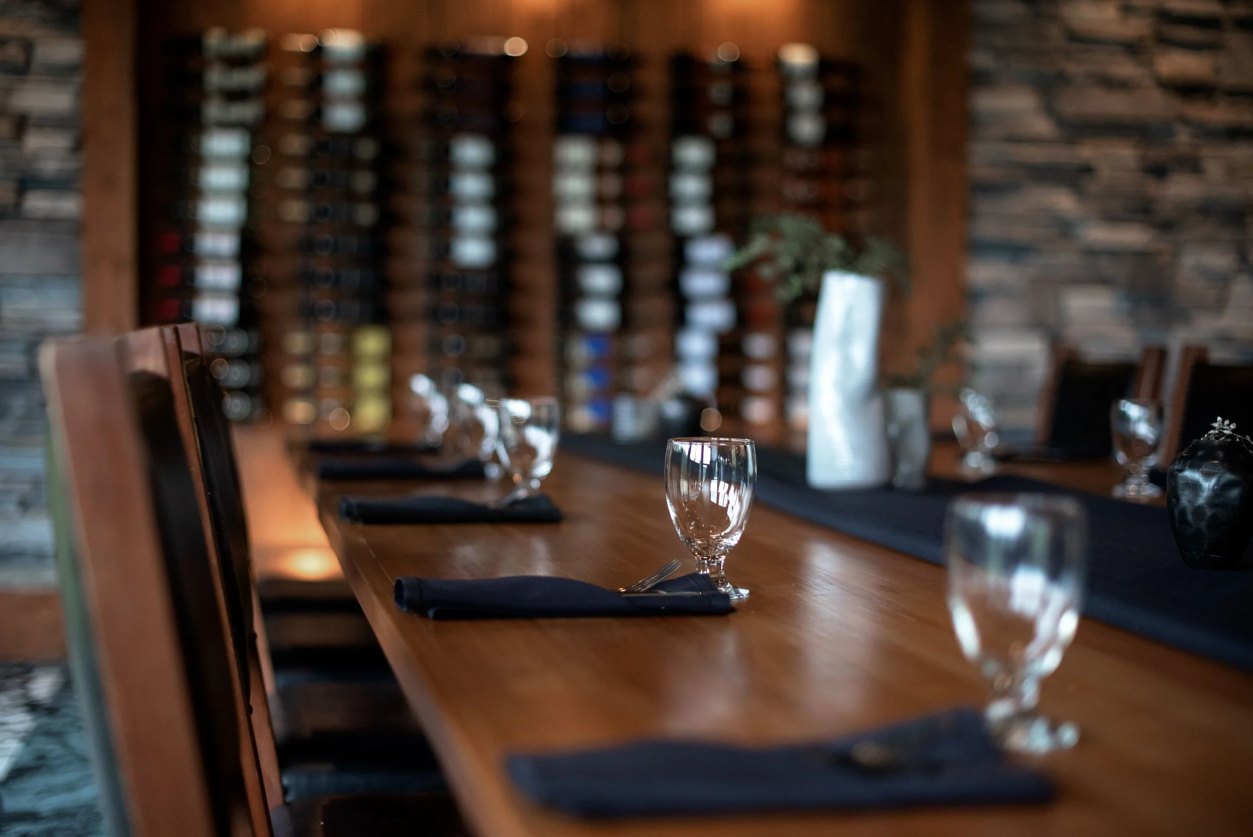 A dining table set with black napkins, a wine glass, and a water glass in a restaurant with wooden walls and a large wine rack in the background.