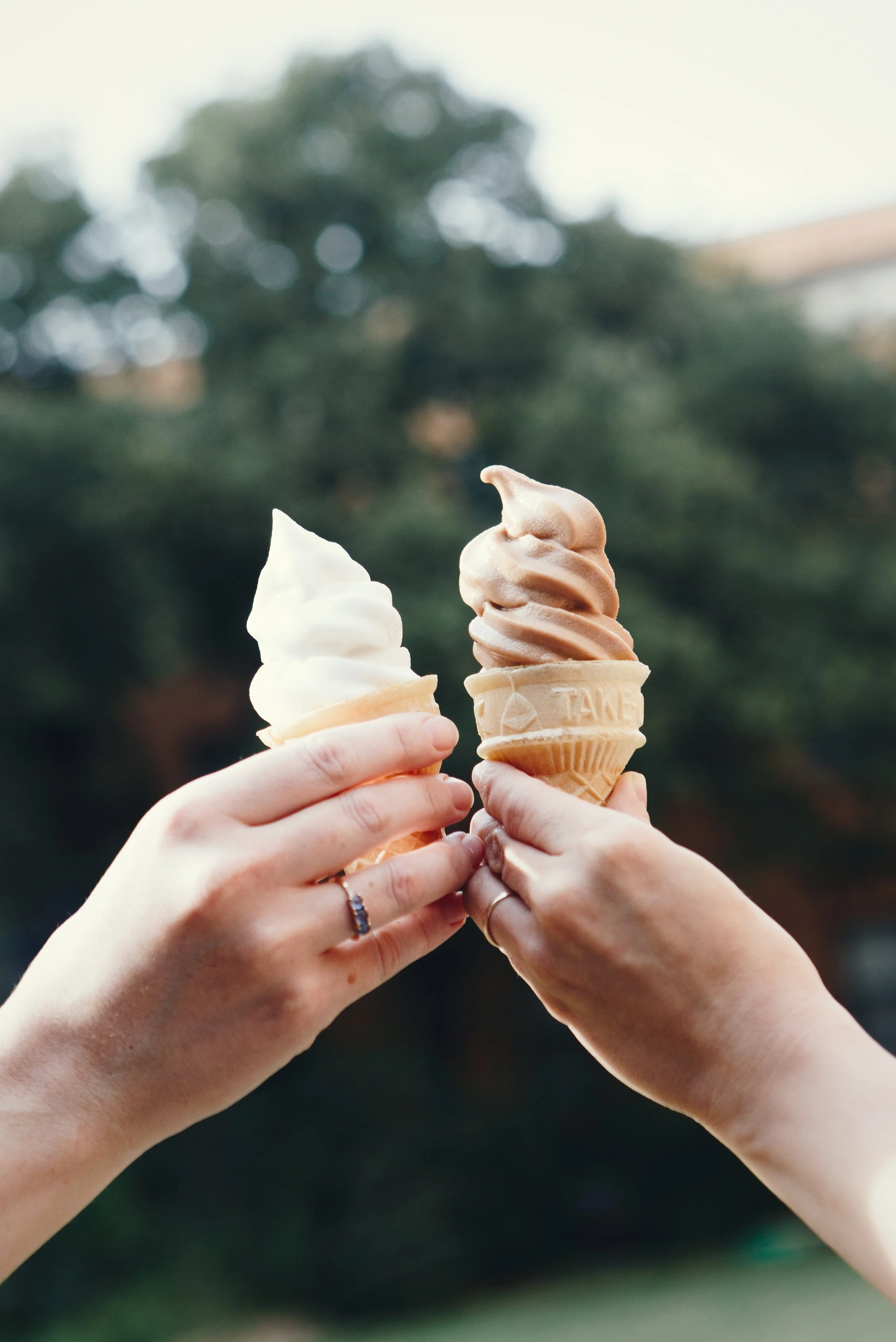 Two hands holding ice cream cones, one with vanilla and the other with caramel-flavored ice cream, outdoors against a blurred background of trees.