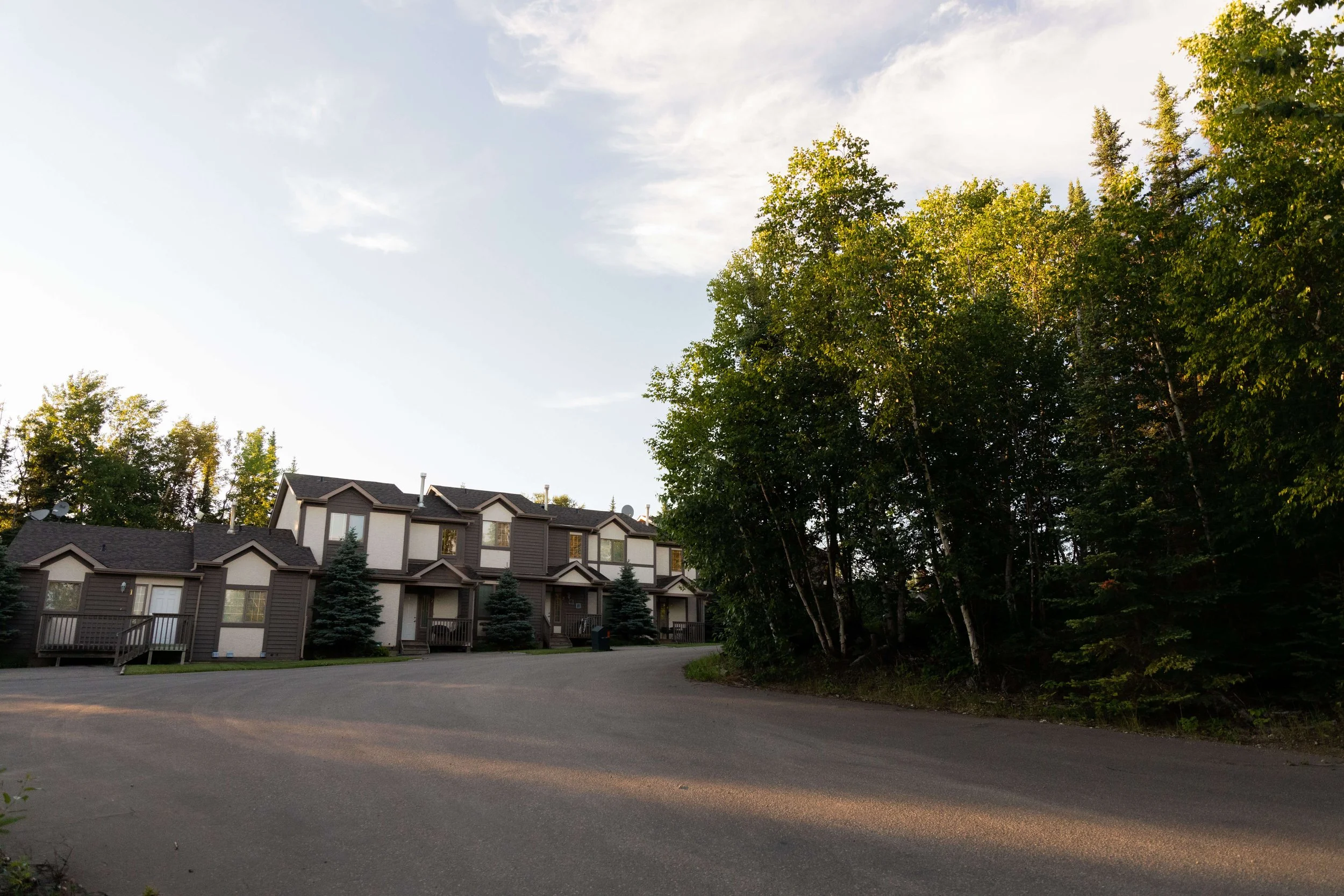 A row of modern townhouses with multiple stories, gray and white exterior, and front porches, surrounded by tall green trees under a partly cloudy sky in a suburban neighborhood.