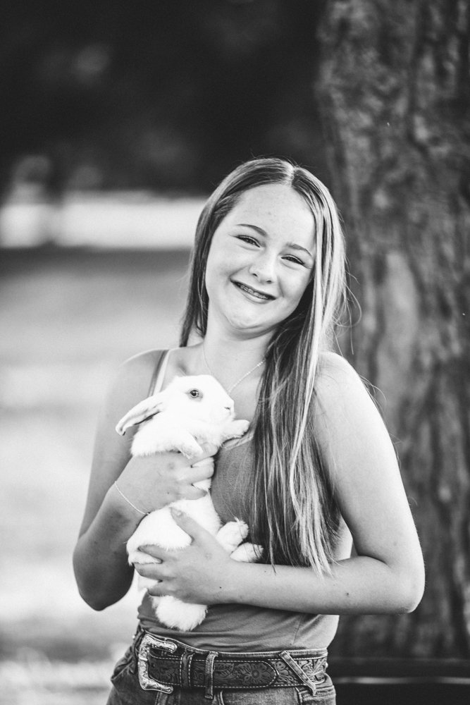 A smiling young woman holding a white rabbit outdoors near a tree in a park, black and white photograph.