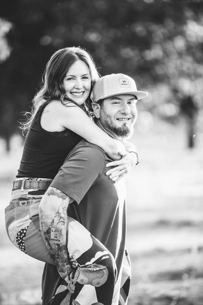 A smiling young woman is piggybacking on a young man in a park with trees, both are smiling, and she has her arms around his shoulders. The photo is in black and white.
