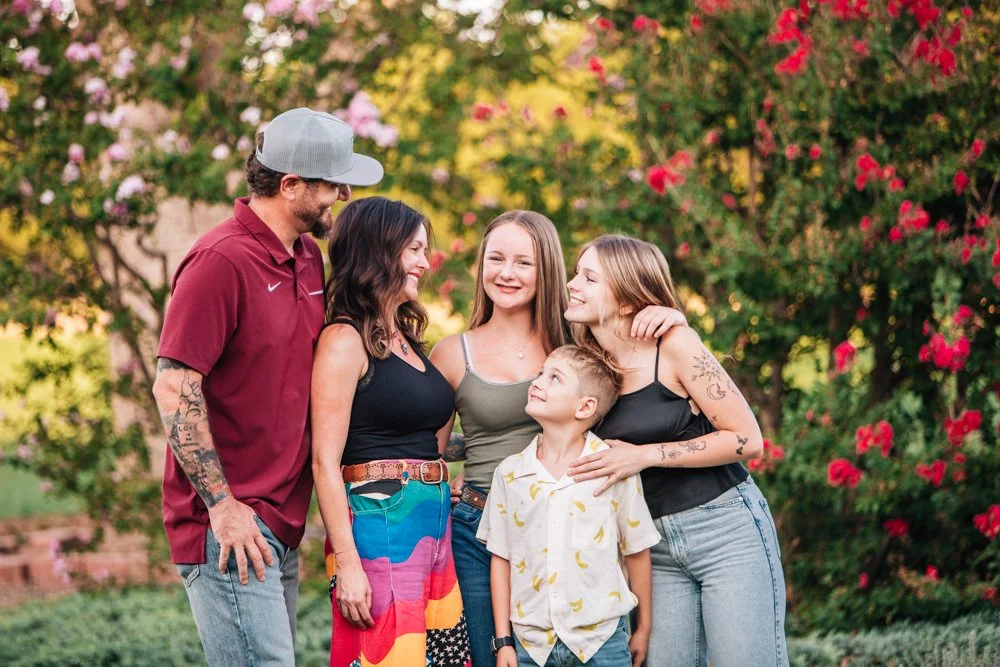 A family of five standing together outdoors in front of flowering bushes, smiling and looking at each other.