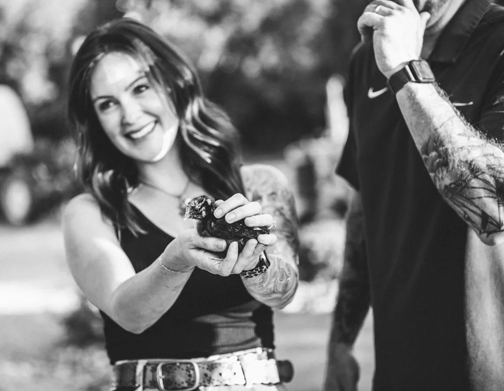 A smiling woman with wavy hair holding a small puppy, with a tattooed man standing nearby, outdoors in black and white.