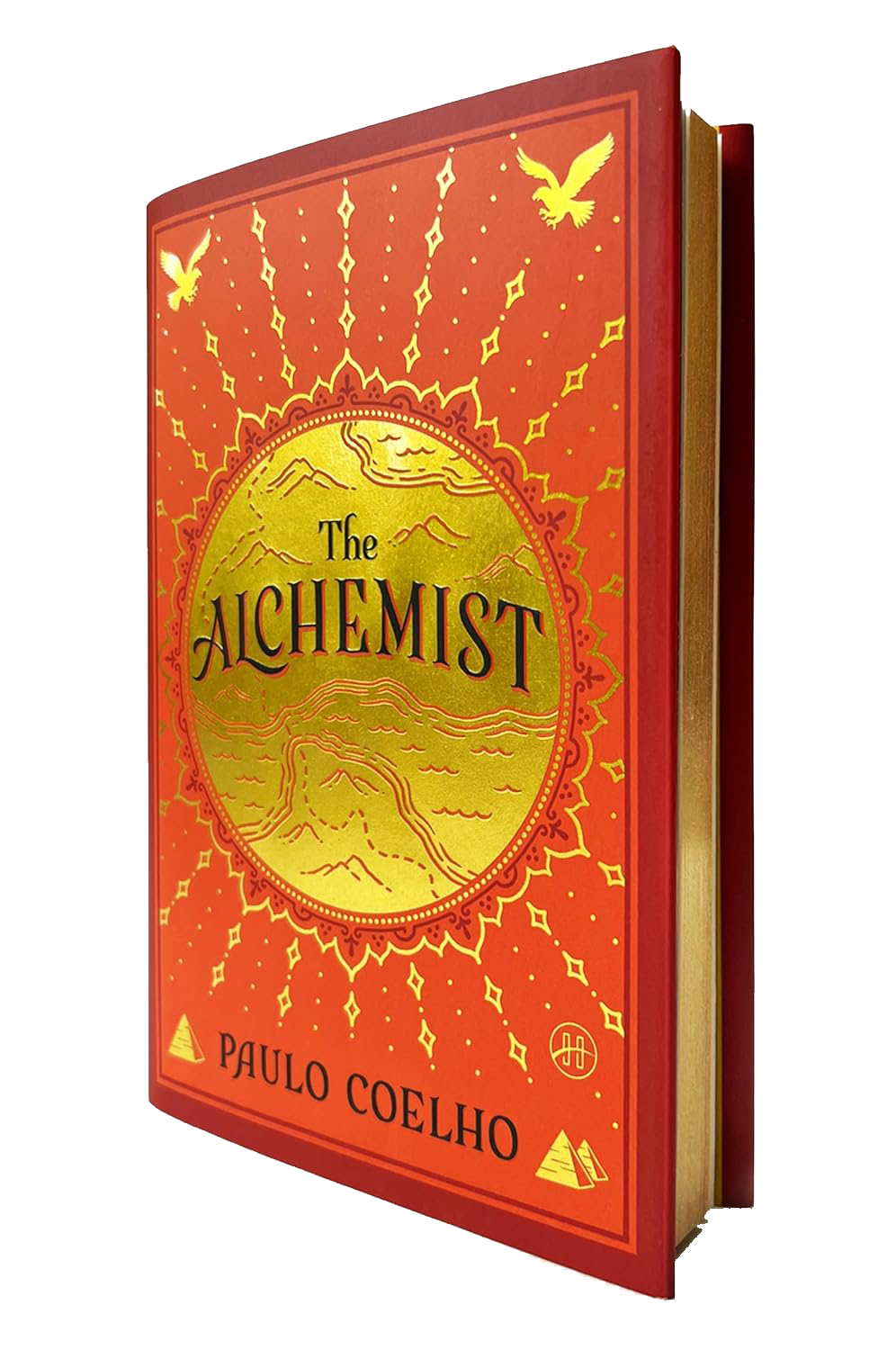 The book titled "The Alchemist" by Paulo Coelho, with an orange and yellow cover featuring desert and mountain illustrations, surrounded by decorative patterns and symbols including pyramids and birds.