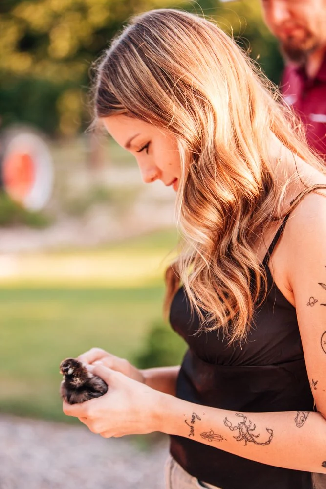 A young woman with long, wavy hair looking down and holding a small chick in her hands outdoors, with a man blurred in the background during sunset.